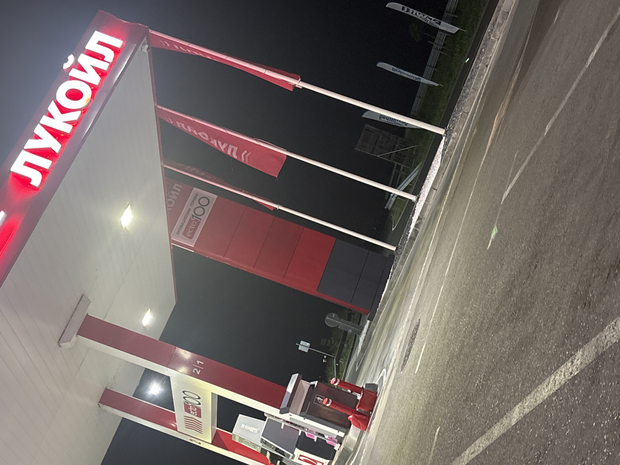 The image shows a Lukoil gas station in Burgas, Bulgaria at night. A bright red "ЛУКОЙЛ" sign is visible at the top of the image, illuminated by white lights. Below the sign, red and white pillars support a canopy over a gas pump area. One of the pillars has a sign that says "ECTO 100" and "2/1". Red flags with white text, also appearing to be Lukoil branding, are planted on poles to the right of the gas station. The ground is asphalt, wet and reflective, with white lane markings. In the background, there is a dark, blurred area that could be the night sky or surrounding landscape. The image is taken from a low angle, looking up towards the gas station. There are no people visible in the image.