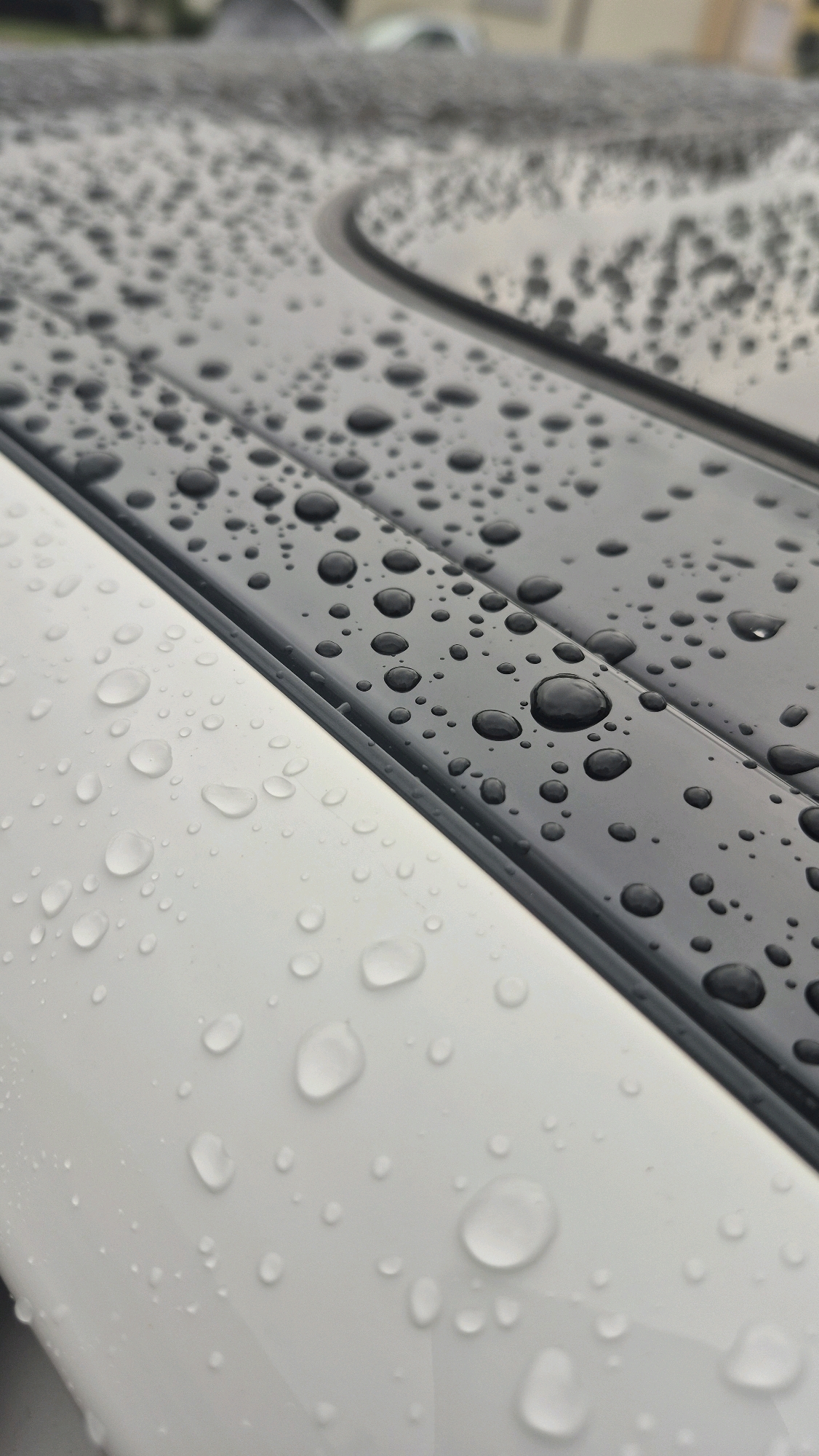 The image shows a close-up, angled view of a car's roof after rainfall, with visible water droplets scattered across both the white painted surface and the black trim. The focus is on the texture and sheen of the water on the car's exterior. The background is blurred, suggesting an outdoor setting, possibly a street or parking lot in Unterensingen, Germany, given the location context. No people or specific landmarks are identifiable.