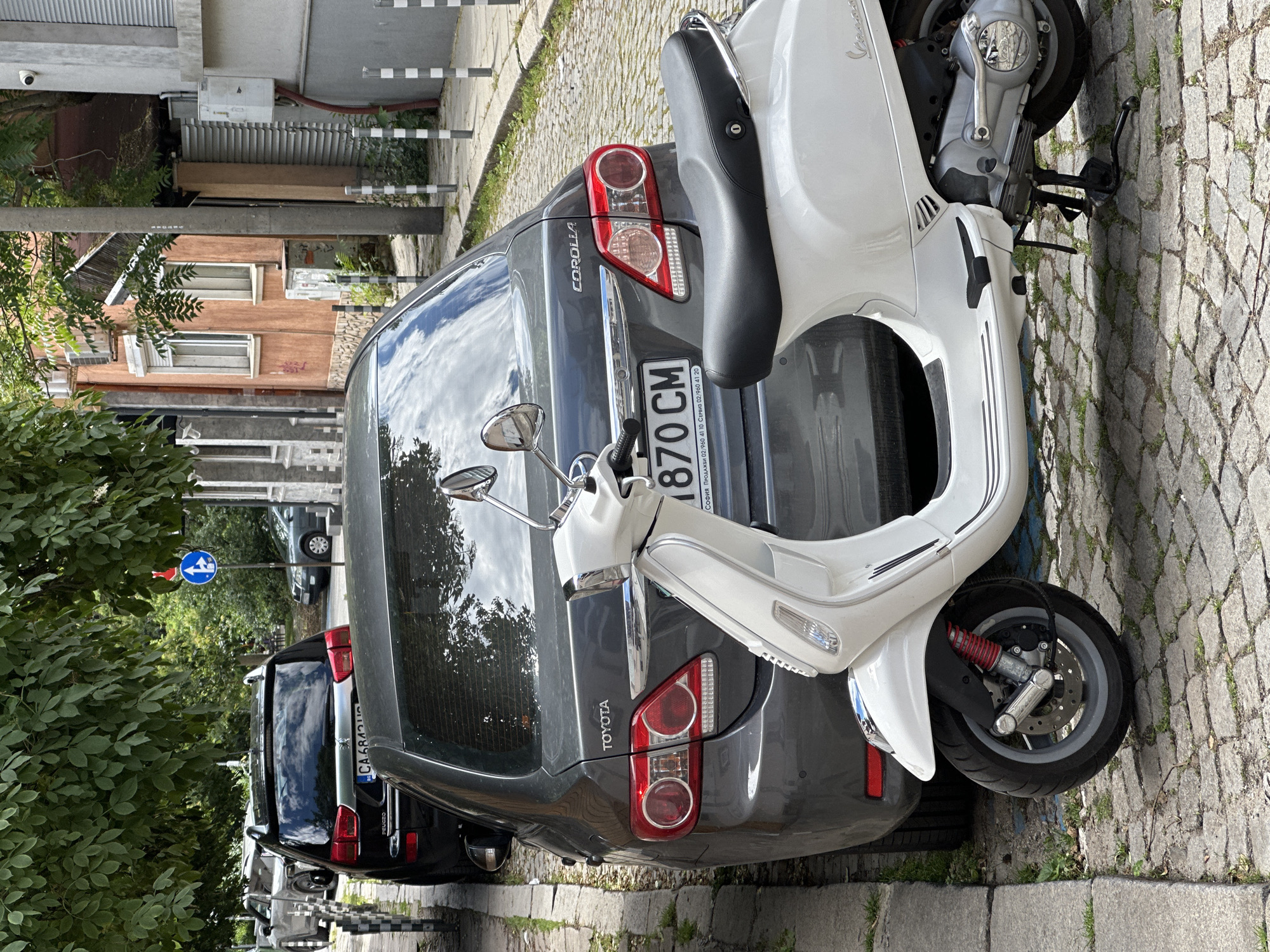 In Sofia, Bulgaria, a white Vespa scooter is parked in front of a dark gray Toyota Corolla on a cobblestone street. The scooter's handlebars, mirrors, front wheel with red suspension, and part of its engine are visible. The Toyota Corolla has its rear end facing the camera, with its taillights, "TOYOTA" and "COROLLA" badging, and a license plate with Cyrillic characters. Behind the Corolla, a black SUV is partially visible. Trees with dense green foliage overhang the scene, casting shadows. The background reveals buildings with orange and gray facades, a traffic sign, and other parked vehicles on the street.
