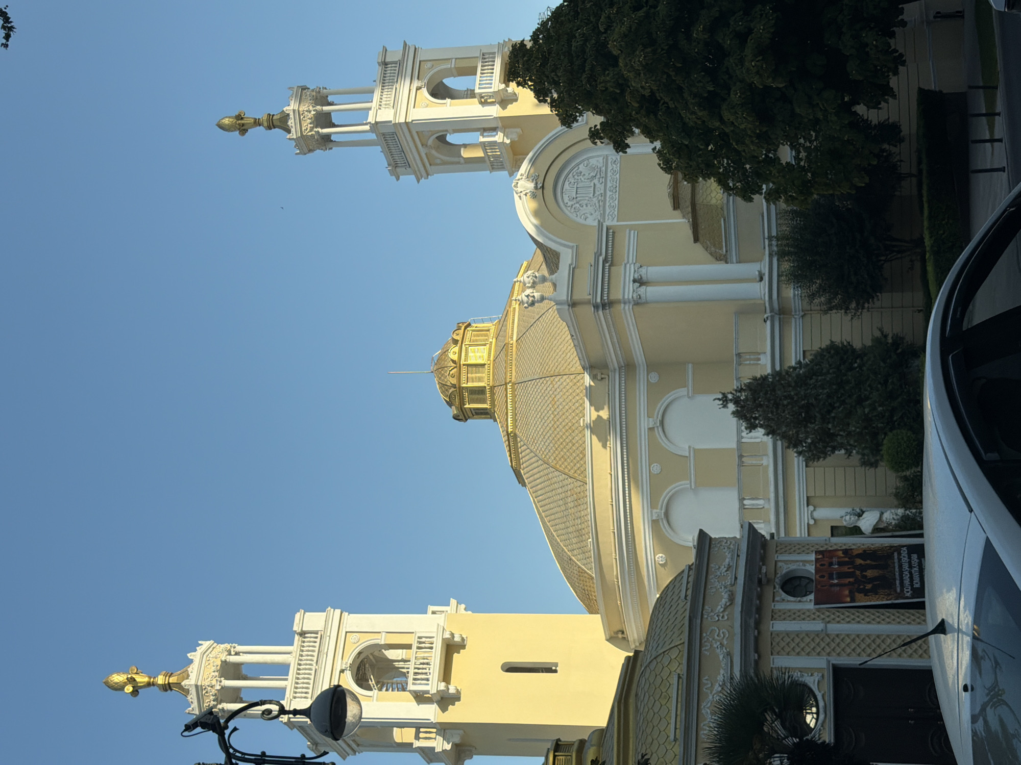The image shows the Azerbaijan State Academic Opera and Ballet Theatre in Baku, Azerbaijan. The facade of the building is a light yellow, with white decorative elements. Two towers flank the central domed structure, each topped with a golden finial. The dome itself is also golden and appears to be covered in scales. There are several trees in the foreground, some of which are dark green and leafy, obscuring parts of the building. A white car is visible in the lower portion of the image, with its roof and windshield taking up a significant area. A poster is displayed on the exterior of a smaller structure to the left of the main building. The sky is a clear, bright blue, suggesting it is daytime.