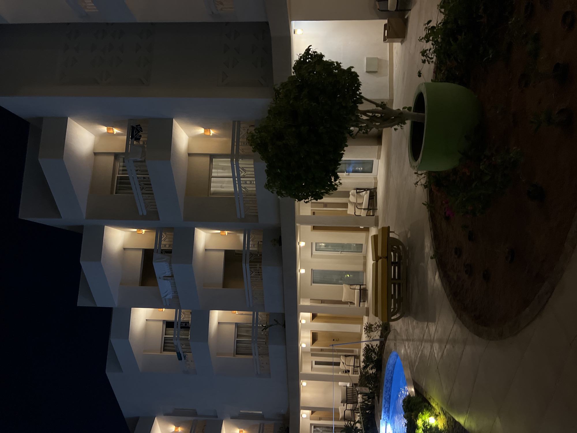 The image depicts a nighttime scene at a resort in Sharm El Sheikh, Egypt. The focus is on the exterior of a multi-story hotel building with balconies illuminated by orange outdoor sconces. In the foreground, a portion of a blue swimming pool is visible on the left, bordered by a paved walkway with embedded yellow lights. A low-lying lounge bed with a yellow cushion is placed on the pavement. A tree in a large green planter sits in the center of the courtyard, surrounded by a garden bed. The ground level of the hotel features several doors and large windows, with some outdoor seating areas visible. The overall ambiance is one of quiet resort hospitality at night.