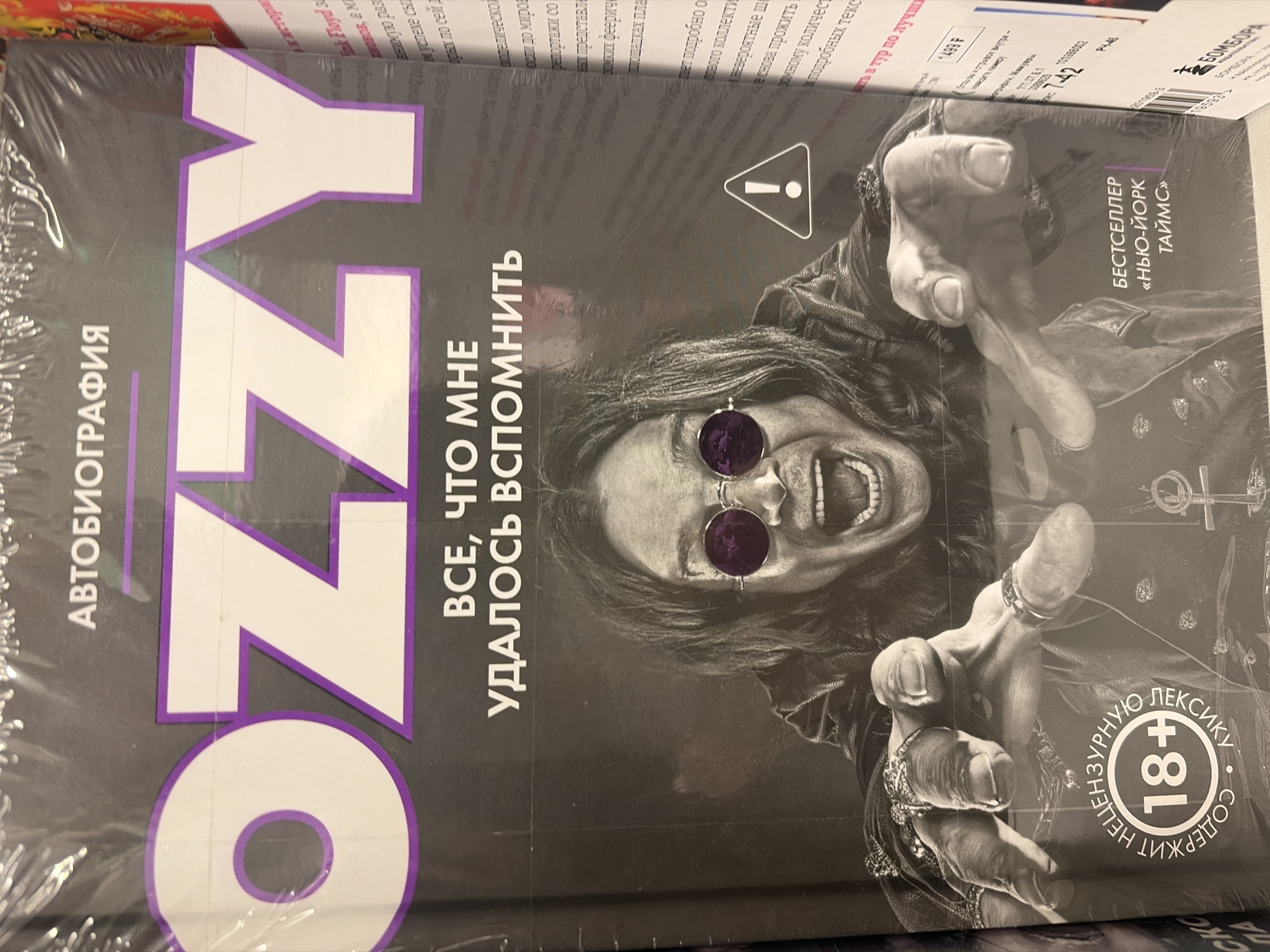 The image displays a shrink-wrapped book titled "OZZY," likely an autobiography, situated in a retail environment such as a bookstore in Saint Petersburg, Russia. The main subject of the book cover is a striking black-and-white portrait of a person resembling Ozzy Osbourne. He is depicted with his arms outstretched towards the viewer, wearing dark, round sunglasses with a purple tint, a leather jacket, and several rings on his fingers. His mouth is wide open, suggesting a scream or laugh, conveying a sense of raw energy and intensity.
The prominent title "OZZY" is rendered in large white letters with a purple outline. To its left, the word "АВТОБИОГРАФИЯ" (Autobiography) is printed vertically. Below the main title, the phrase "ВСЕ, ЧТО МНЕ УДАЛОСЬ ВСПОМНИТЬ" (Everything I Managed to Remember) is displayed.
Notable details on the cover include a warning sign (an exclamation mark in a triangle), a circular emblem indicating "СОДЕРЖИТ НЕЦЕНЗУРНУЮ ЛЕКСИКУ 18+" (Contains uncensored language 18+), and a rectangular label proudly stating "БЕСТСЕЛЛЕР «НЬЮ-ЙОРК ТАЙМС»" (Bestseller "New York Times"). An ankh symbol is also visible on the right side of the cover.
The book is encased in a transparent plastic wrap, indicating it is new and for sale. In the blurred background, other books and shelving units, along with various price tags and product labels, are discernible, reinforcing the setting of a retail store. Some visible text in the background, like "1499 P" (rubles) and company names like "БОМБОРА," further confirm the commercial context in Russia.