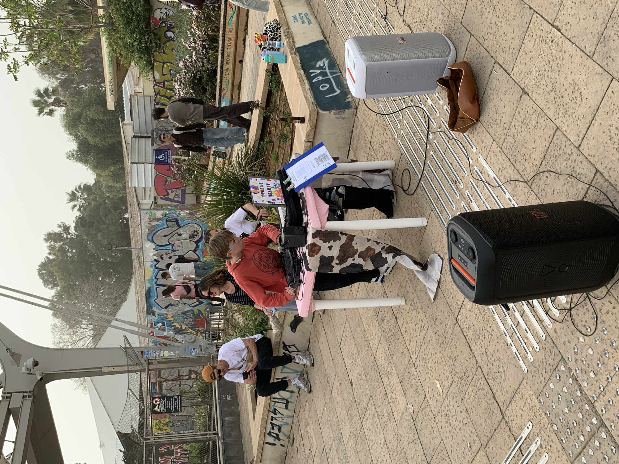 The image shows a scene in Tel Aviv, Israel. A group of people are gathered around a makeshift table set up on a tiled public walkway. The table has a cow-print cloth and a pink covering, and there's a sign with "@PUBLIC TRANCE" on it. Several people are seated or crouching near the table, including one in a red sweatshirt and another in a white shirt. Two JBL speakers are positioned nearby, one white and one black. A brown bag is also present near the white speaker. A security camera is mounted atop a metal beam in the background, and a wall with graffiti is visible behind the group. Other people are walking in the background as well.
