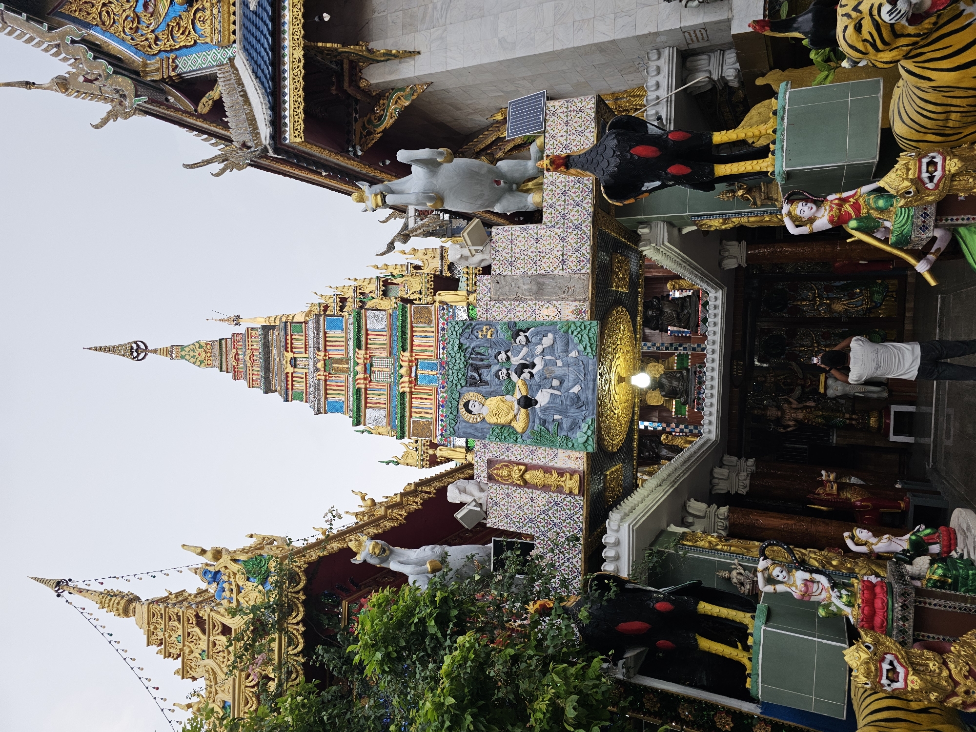 This is a low-angle, wide shot of Wat Samphran, a unique temple located in Nakhon Pathom Province, west of Bangkok, Thailand. The temple is most famous for its giant, pink dragon-wrapped tower. The scene captures the elaborate facade of the temple, which is adorned with intricate golden decorations, colorful mosaics, and numerous statues.
In the foreground, on the left, is a large, golden statue of a tiger's head. To the right of the tiger, a statue of a female figure in a green and red outfit is positioned on a lotus pedestal, holding a golden spear and entwined with a snake. Behind her, a statue of a rooster with red markings stands prominently on a green platform. Further back, more statues of deities and mythical creatures are visible, including what appears to be an elephant statue to the right.
The central part of the temple features a tiered, pyramid-like structure, also adorned with gold and colorful mosaics, topped with a spire. A relief carving depicting Buddha in meditation with disciples is prominently displayed on the lower section of this central structure. Flanking this structure are two large, grey statues of mythical horse-like creatures, one on each side, standing on elevated platforms.
The upper portions of the temple show intricate golden roof designs with finials. A single solar panel is visible on the right side of the central structure.
A person dressed in a white t-shirt and dark pants is standing in front of the temple entrance, holding up a phone to take a photograph. The background is a slightly overcast sky. The overall impression is of a vibrant, visually rich religious site with a strong emphasis on ornate decoration and figurative art.