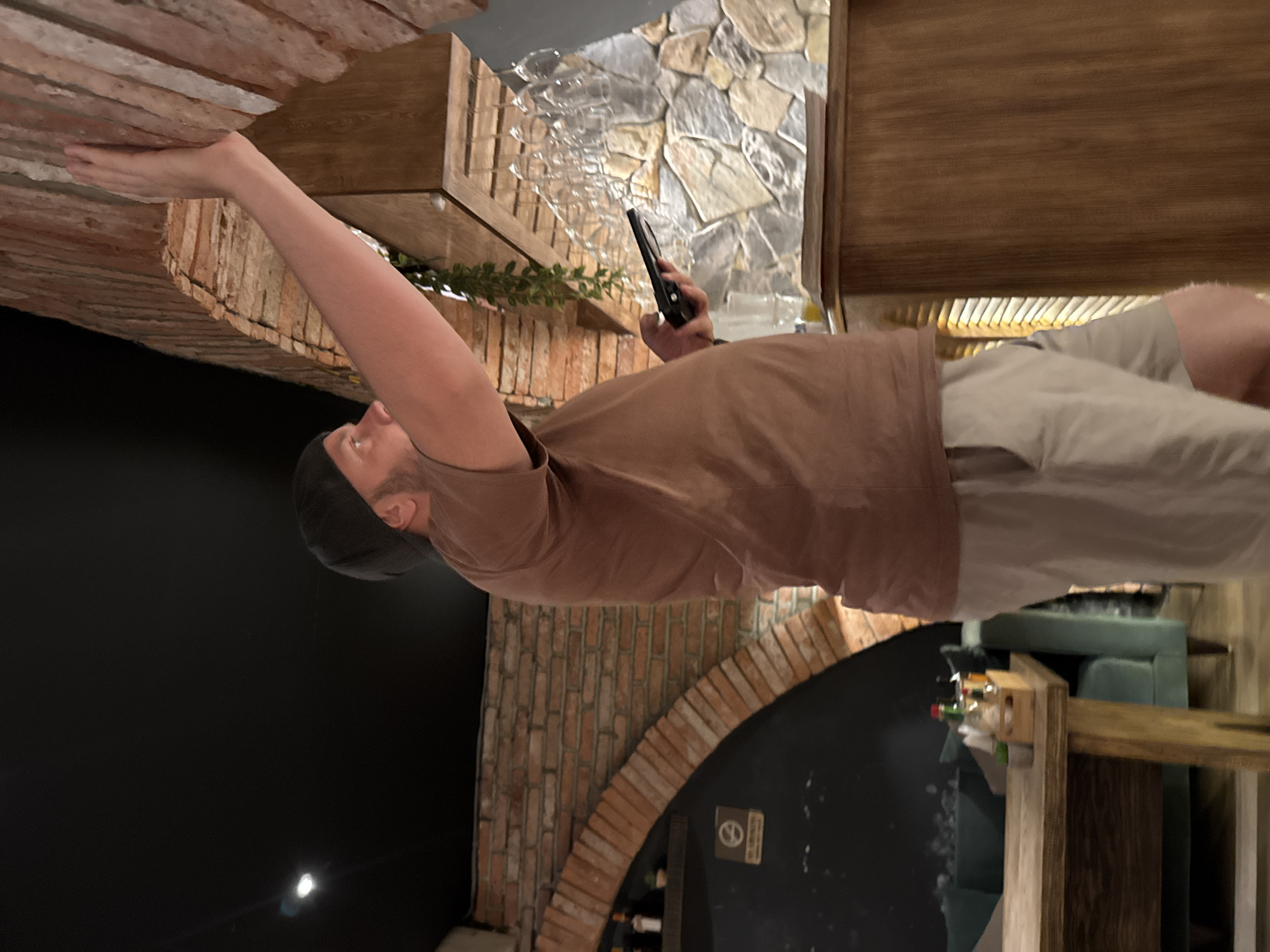 A man wearing a brown t-shirt and light-colored shorts and a black baseball cap is standing and reaching up with his right hand to touch a brick wall. He is holding a black mobile phone in his left hand. The man is standing in what appears to be a restaurant or bar, with brick arches and stone walls visible in the background. There are wine glasses hanging from a rack above a wooden counter on the right side of the image. A wooden table with a green couch behind it is visible on the left side of the image. The lighting is dim, suggesting it is evening. The specific location in Hanoi, Vietnam cannot be determined from the image alone.