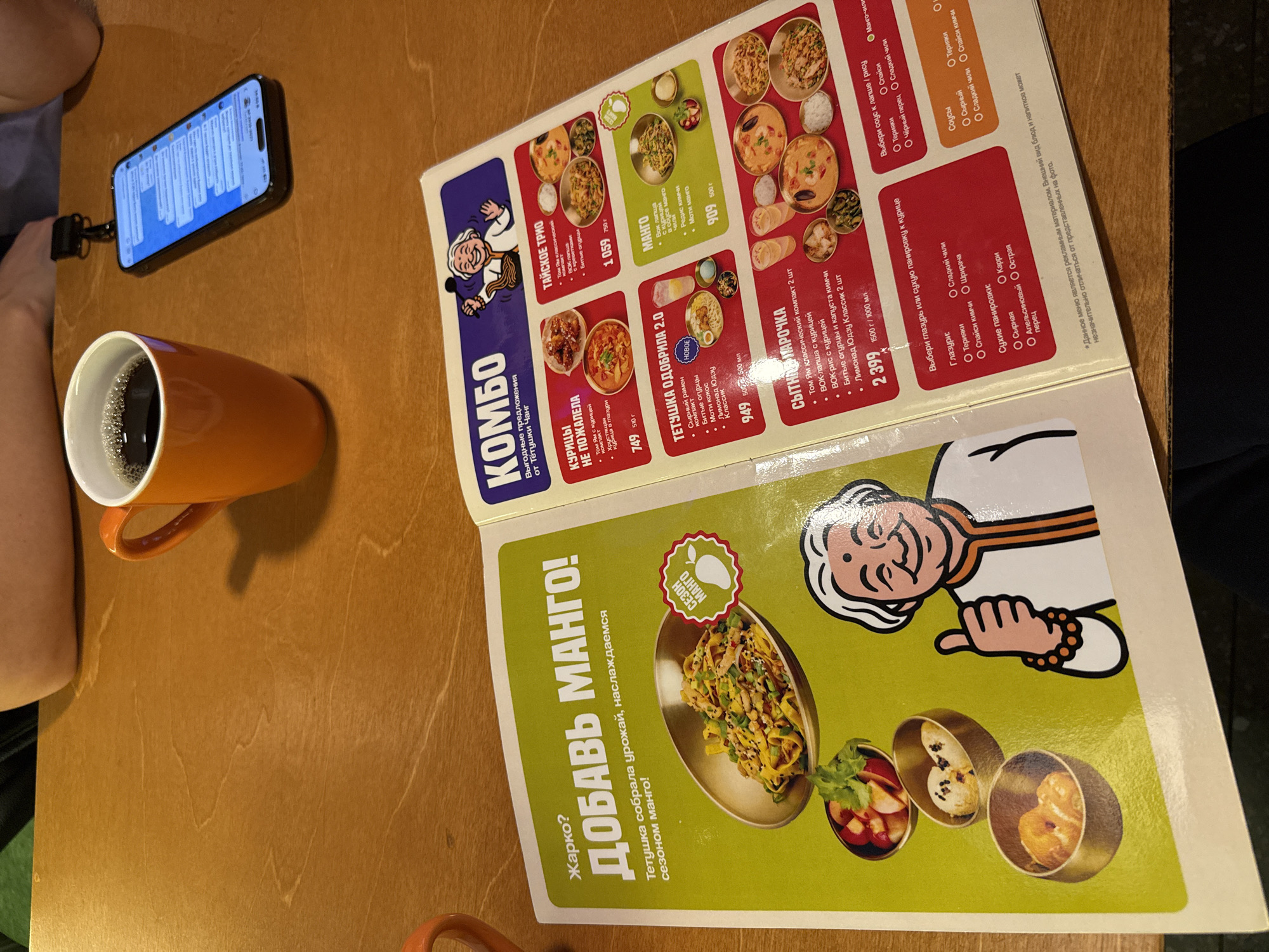 The image depicts a close-up view of a table in a restaurant setting. On the table, there are two orange mugs, one filled with a dark liquid, possibly coffee, and an open smartphone displaying a chat interface. To the right of the mugs, a menu is open, featuring colorful illustrations and text. The menu appears to be for an Asian restaurant, with sections like "КОМБО," "КУРИЦЫ НЕ ПОЖАЛЕЛА," and "ТЕТУШКА ОДОБРИЛА 2.0." The menu details various food options, including dishes with chicken, noodles, and mango. The background shows parts of a person's arm and shoulder, suggesting they are seated at the table. The overall environment suggests a casual dining experience. The presence of the menu and the specific dish descriptions indicate a restaurant located in Izhevsk, Russia, where this particular establishment is situated.
