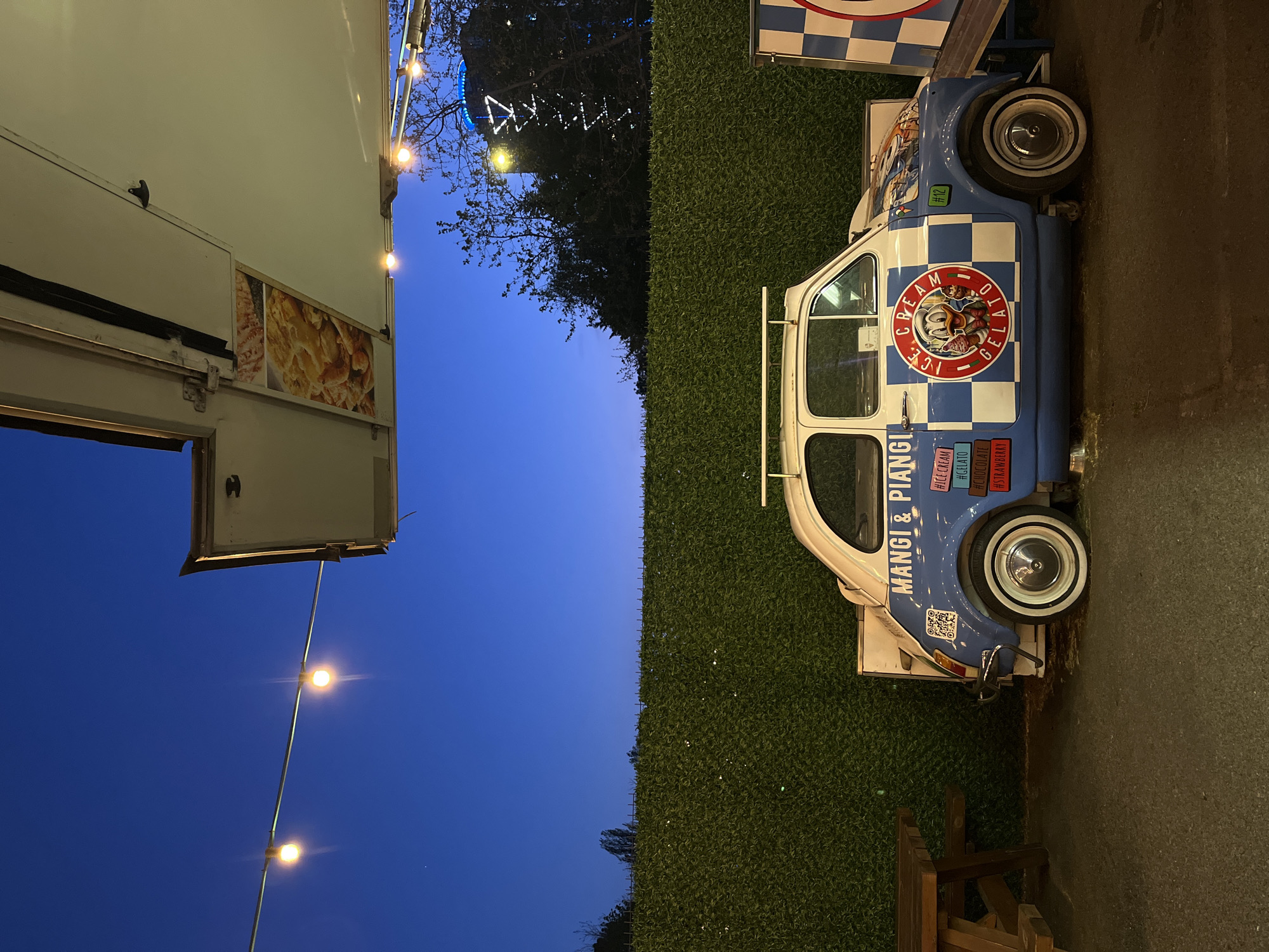 The image displays an outdoor food and dessert vending scene at dusk or night, illuminated by artificial string lights. The deep blue twilight sky serves as the backdrop for the setup.
On the left side, a large, white food truck or trailer is visible with its serving hatch open. Inside the opening, a photographic display shows a close-up of a savory food item, possibly pizza or focaccia. Several warm yellow string lights are suspended horizontally in front of the truck.
To the right of the food truck, separated by a dark green, textured artificial hedge, stands a distinctive blue and white checkered microcar, repurposed as an "ICE CREAM GELATO" vendor. This vehicle, resembling a classic Italian microcar like a Fiat 500 or Piaggio Ape, features white-wall tires.
The side of the microcar is adorned with several details:
* A circular logo reads "ICE CREAM GELATO" surrounding a cartoon character, likely Donald Duck, within a red circle, with "1921" subtly included.
* Below the logo, the Italian phrase "MANGI & PIANGI" (Eat & Cry) is prominently displayed.
* A vertical list of hashtags indicates offerings: "#ICE CREAM", "#GELATO", "#CHOCOLATE", and "#STRAWBERRY".
* A QR code is also visible on the side of the vehicle, along with a small green sticker marked "#12".
Tree branches are visible against the twilight sky in the background, suggesting an open-air environment. The ground beneath the vehicles and hedge is a dark, paved surface, with a part of a wooden bench or table visible in the bottom right corner. No people are present, but the vibrant setup indicates a lively commercial activity, likely in Sofia, Bulgaria.