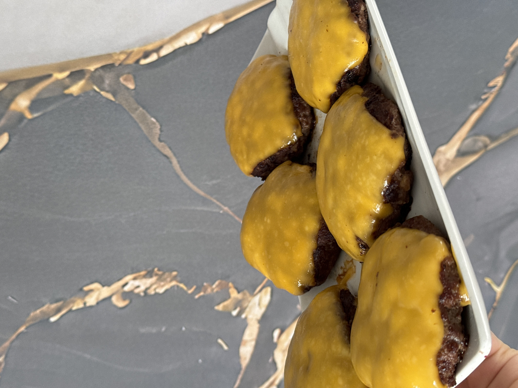 The media file shows a close-up, top-down view of several cheeseburger patties arranged in a white rectangular dish. The patties are dark brown and covered with melted yellow cheese, which drips down the sides. The dish is placed on a surface with a dark grey background featuring prominent, abstract gold and black marbling patterns. The lighting appears to be natural, suggesting an indoor setting with diffused light. There are no people visible in the frame. The focus is on the food item, highlighting the texture of the patties and the melted cheese. The overall presentation is simple, with the food being the central subject.