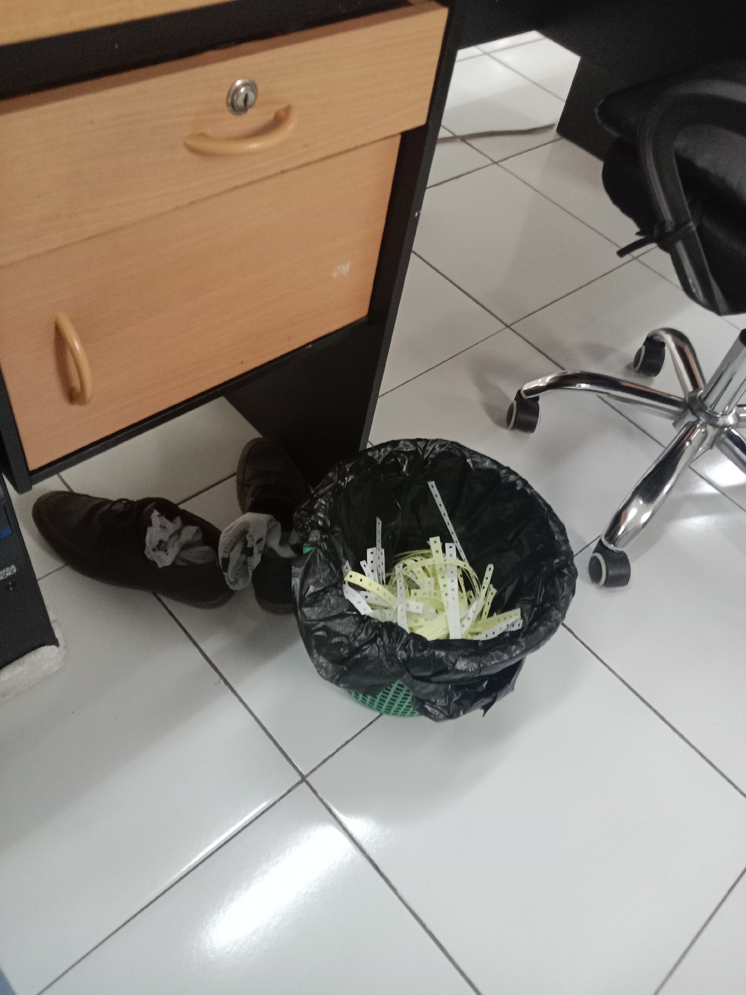 In a brightly lit room with white tiled floors, a black plastic trash bin filled with shredded white paper sits on a green plastic grate. Next to it, a pair of dark brown leather shoes, each with a white sock partially stuffed inside, rests on the floor. In the background, a wooden filing cabinet with a silver lock and a curved handle is visible, and the chrome base of an office chair with black wheels is also present. The scene suggests a typical office environment, potentially in Semarang, Indonesia, where an individual has removed their shoes and socks, and discarded paper.