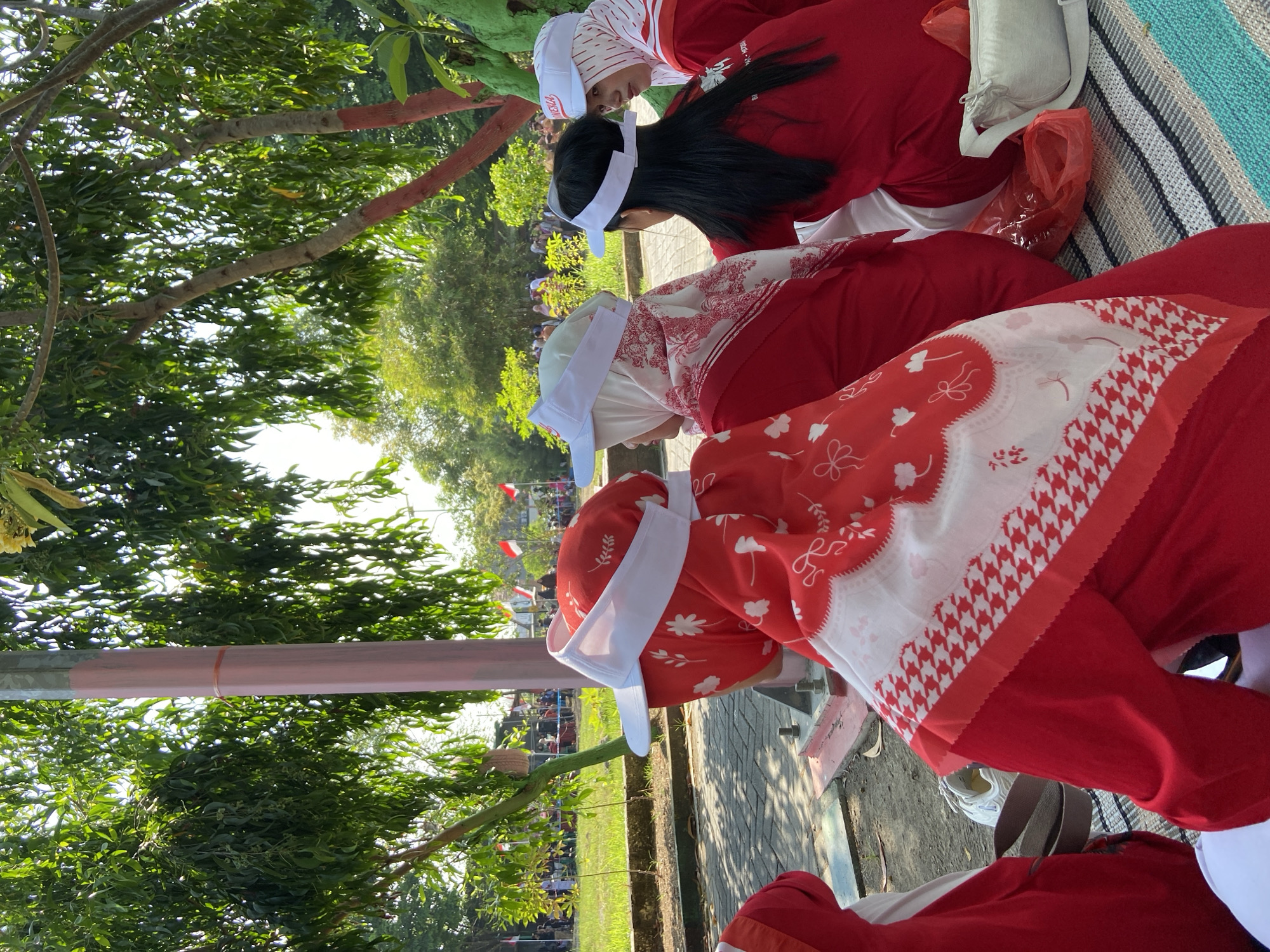 In Gelam, Indonesia, a group of young women, likely students, are seated on a striped mat outdoors, facing away from the camera. They are dressed in matching bright red outfits, with white headscarves and white visors. The women appear to be watching an event, as suggested by the flags in the background. The environment is a park-like setting with trees providing shade. The lighting indicates it is daytime, with sunlight filtering through the leaves. There are other people and flags visible in the distance, suggesting a public gathering or celebration.