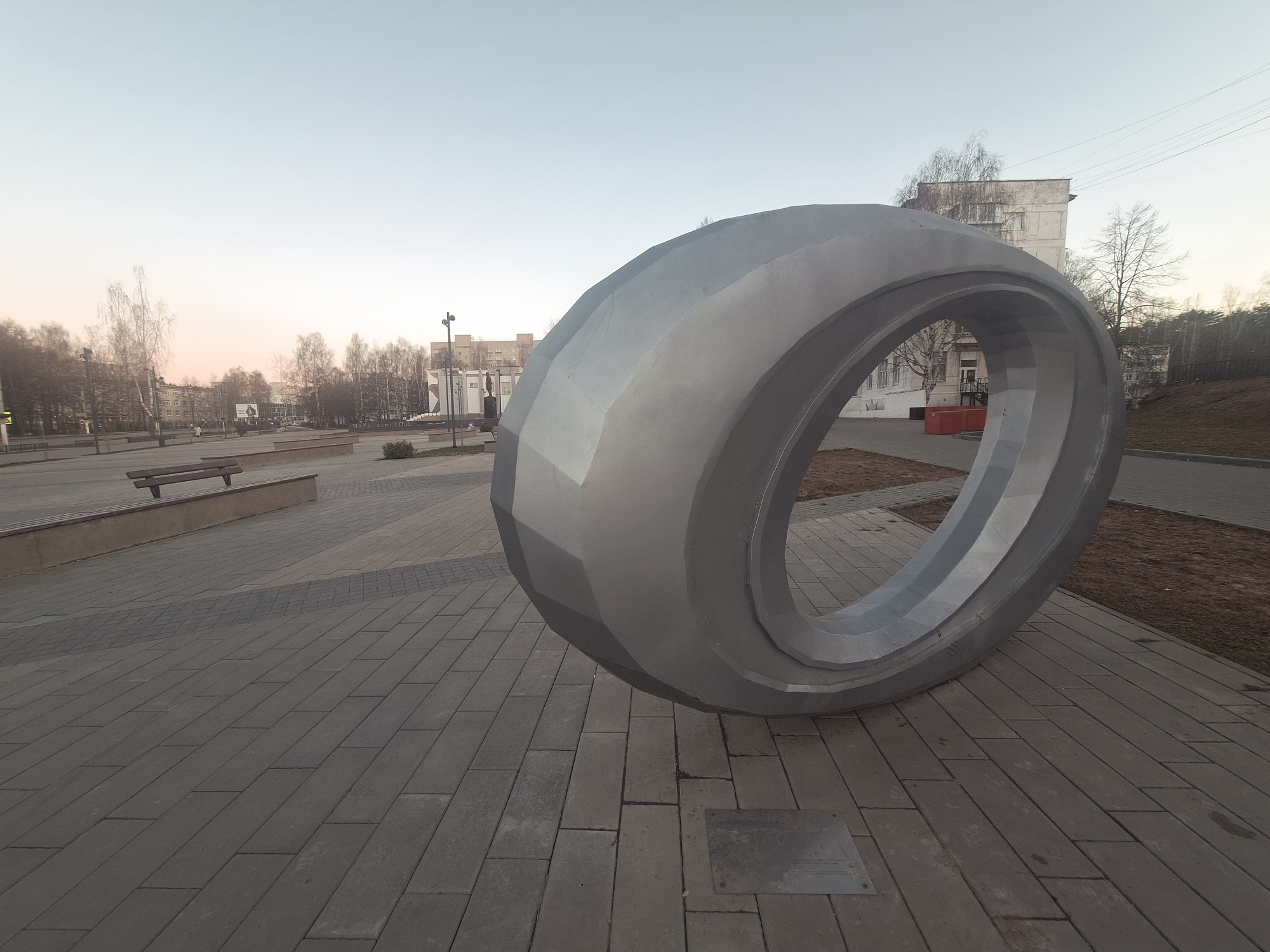 The image depicts a large, silver, torus-shaped sculpture situated in a paved public space. The sculpture has a faceted exterior, giving it a somewhat polygonal appearance. It is positioned on a rectangular paved area that extends into the background. To the left of the sculpture, there's a paved pathway with a bench. In the background, there are trees and buildings. The sky is light, suggesting it's either dawn or dusk. The location is the city of Unknown, Unknown. There are no people visible in the image.