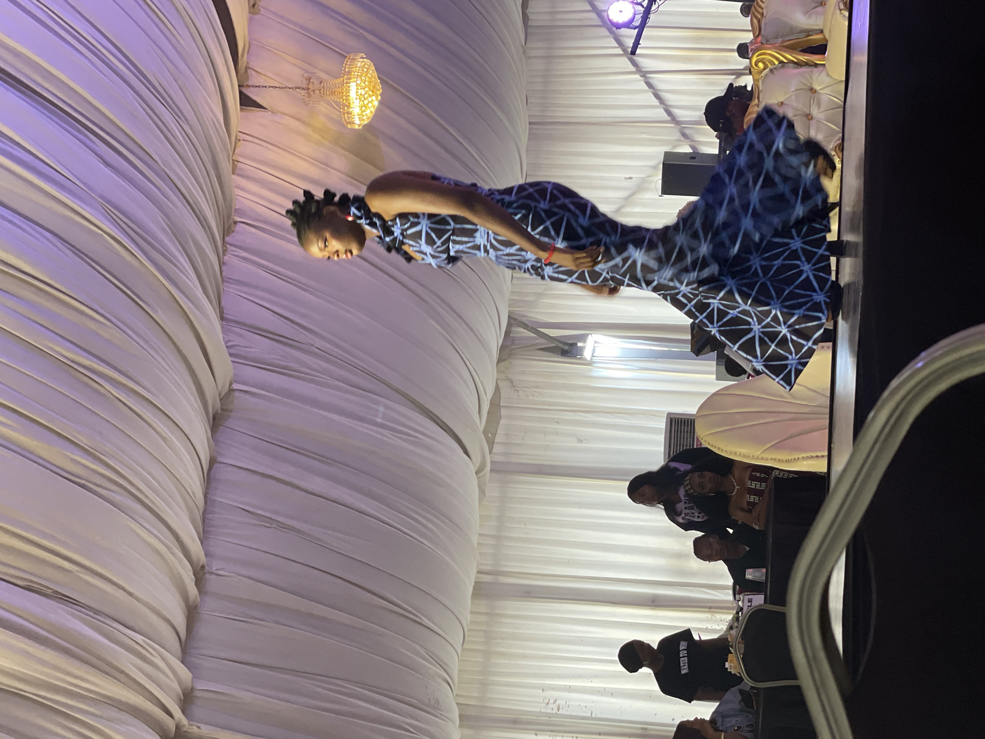 A fashion show is taking place under a draped white tent in Barakin Lerawi, Nigeria. The main subject is a Black female model walking down a runway in a long, fitted blue and black patterned dress that flares out at the bottom. She has her hair styled in braids and wears red earrings. In the background, several people are seated at tables, and a chandelier hangs from the tent ceiling. Other individuals, including staff and audience members, are visible in the middle ground. The environment appears to be an indoor event space, possibly a banquet hall, set up for a fashion presentation.