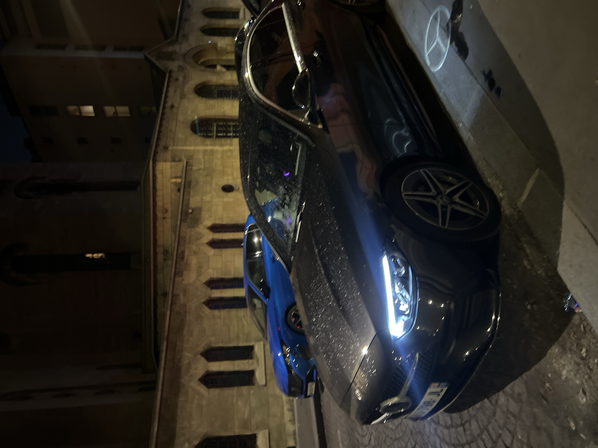 In the city of Paris, France, at night, two cars are parked on a cobblestone street next to a building with Gothic-style architecture. A dark-colored Mercedes-Benz sedan is in the foreground, its headlights on, casting light on the wet street. The car has a license plate that reads "798 JL." In the background, a bright blue Peugeot hatchback is parked. The building behind the cars is illuminated by streetlights, revealing arched windows. The scene suggests a rainy or damp night, as indicated by the reflections and water droplets on the cars and street.