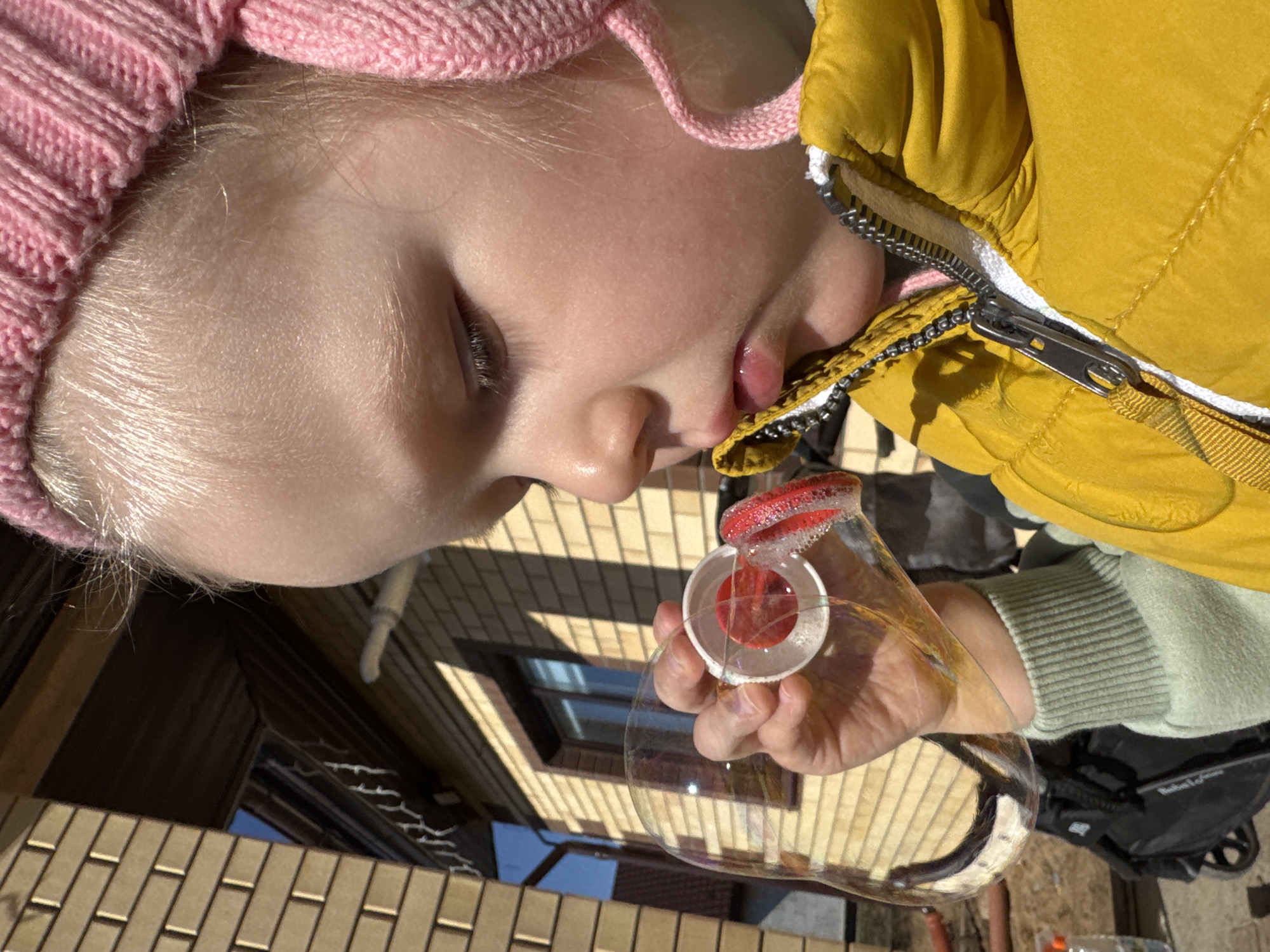 A young child wearing a pink knitted hat and a yellow jacket is holding a bubble wand and blowing bubbles. The background shows a building with brickwork and a window. The image appears to have been taken outdoors, likely in Gornyi (Dvurechenskoe S/P), Russia. A portion of a stroller is visible in the lower right of the frame.