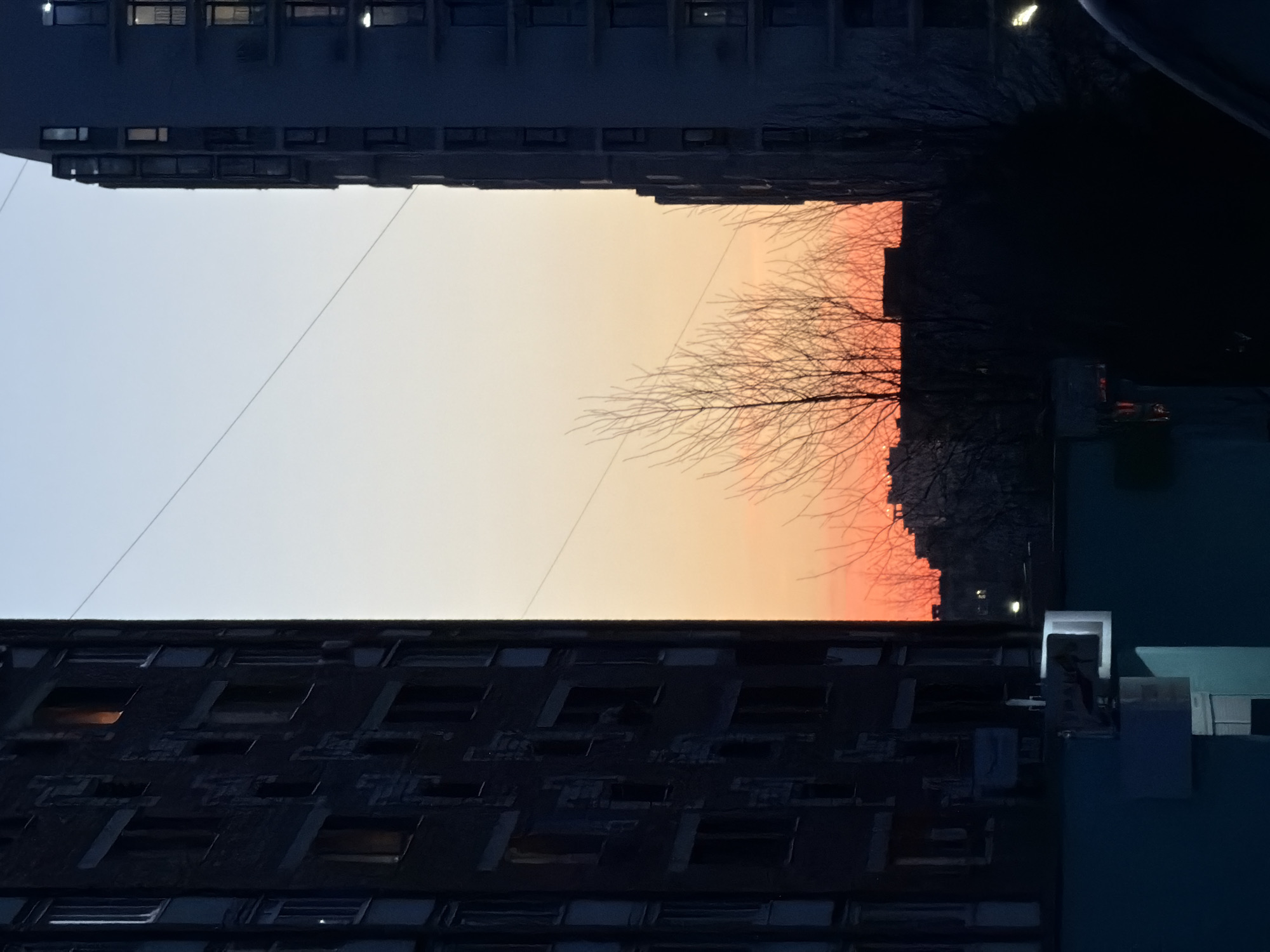 The image captures a view between buildings, possibly in Izhevsk, Russia, given the request's context. The scene depicts a sunset with orange and yellow hues visible between the structures. Bare tree branches are silhouetted against the colorful sky. The buildings appear to be multi-story residential or commercial structures, with numerous windows and a dark, possibly brick or concrete, exterior. A power line stretches across the sky between the buildings. The bottom building appears to have some sort of blue or turquoise exterior paint on a portion of its surface. The image is taken from a low angle, looking up between the buildings.