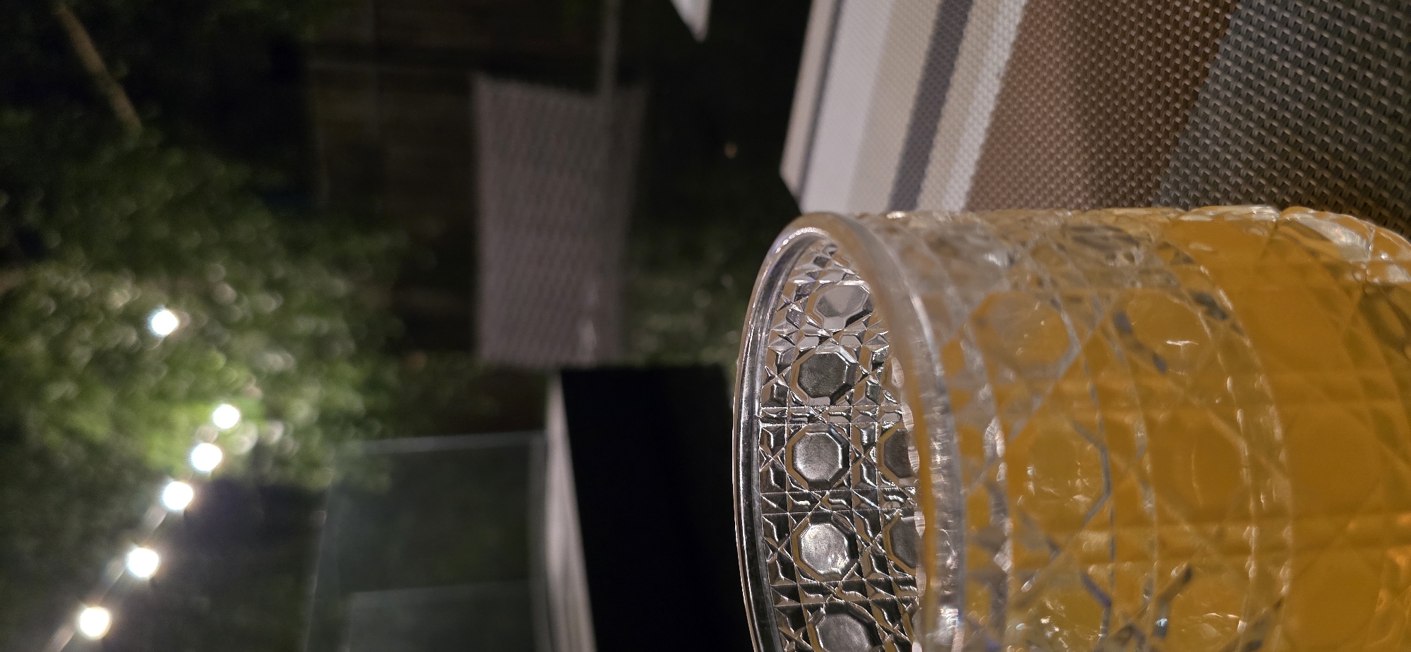 The image depicts an outdoor setting at night, possibly a backyard or patio. In the foreground, a glass filled with an orange liquid, likely juice or a cocktail, sits on a table. The glass has a distinctive textured pattern. Behind the glass, the background is blurred, showing string lights illuminating trees and foliage. A dark, reflective surface, possibly a table or a pool, is visible. In the mid-ground, there appears to be outdoor furniture, a dark rectangular object that could be a grill or a counter, and further back, a fence. The overall ambiance suggests a relaxed evening setting. The image does not contain any specific details that would allow for its location to be identified as being in the city of Kaliningrad, Russia.