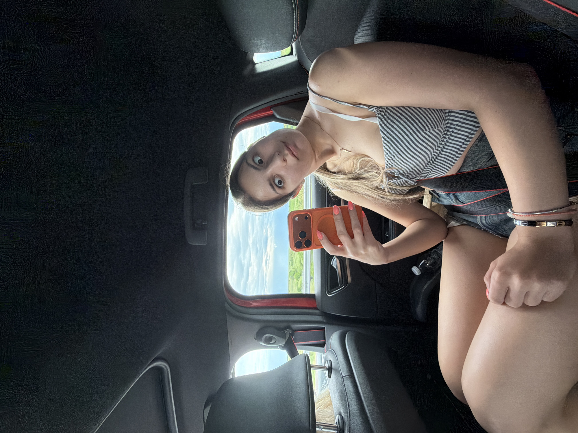 A young woman is sitting in the back seat of a car, holding an orange-cased smartphone in her hand and facing the camera. She is wearing a striped top and denim shorts, and a seatbelt is fastened across her chest. The car's interior is primarily black, with red accents on the seats and doors. Outside the window, the landscape is visible, consisting of a highway and green fields under a partly cloudy sky. There is no explicit evidence to confirm the specific location as Koniovo, Bulgaria.