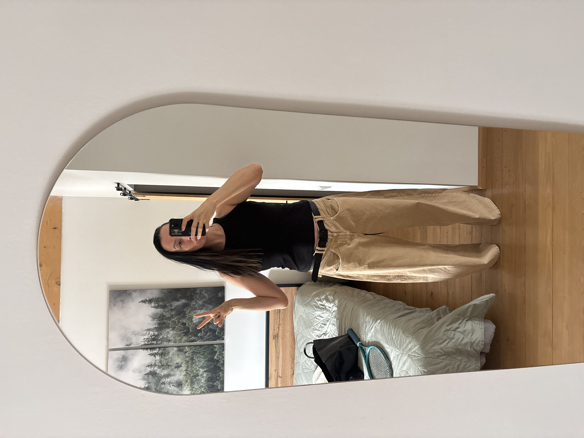 A woman is reflected in a mirror. She is wearing a black tank top and khaki pants, holding a smartphone to take a selfie. She makes a peace sign with her left hand. The mirror reflects a bedroom interior, including a bed with a light green quilt, a tennis racket, a black bag, a wood floor, a wall with a forest-themed artwork, and part of a doorway. The reflection suggests the photograph was taken indoors. The location is unknown.