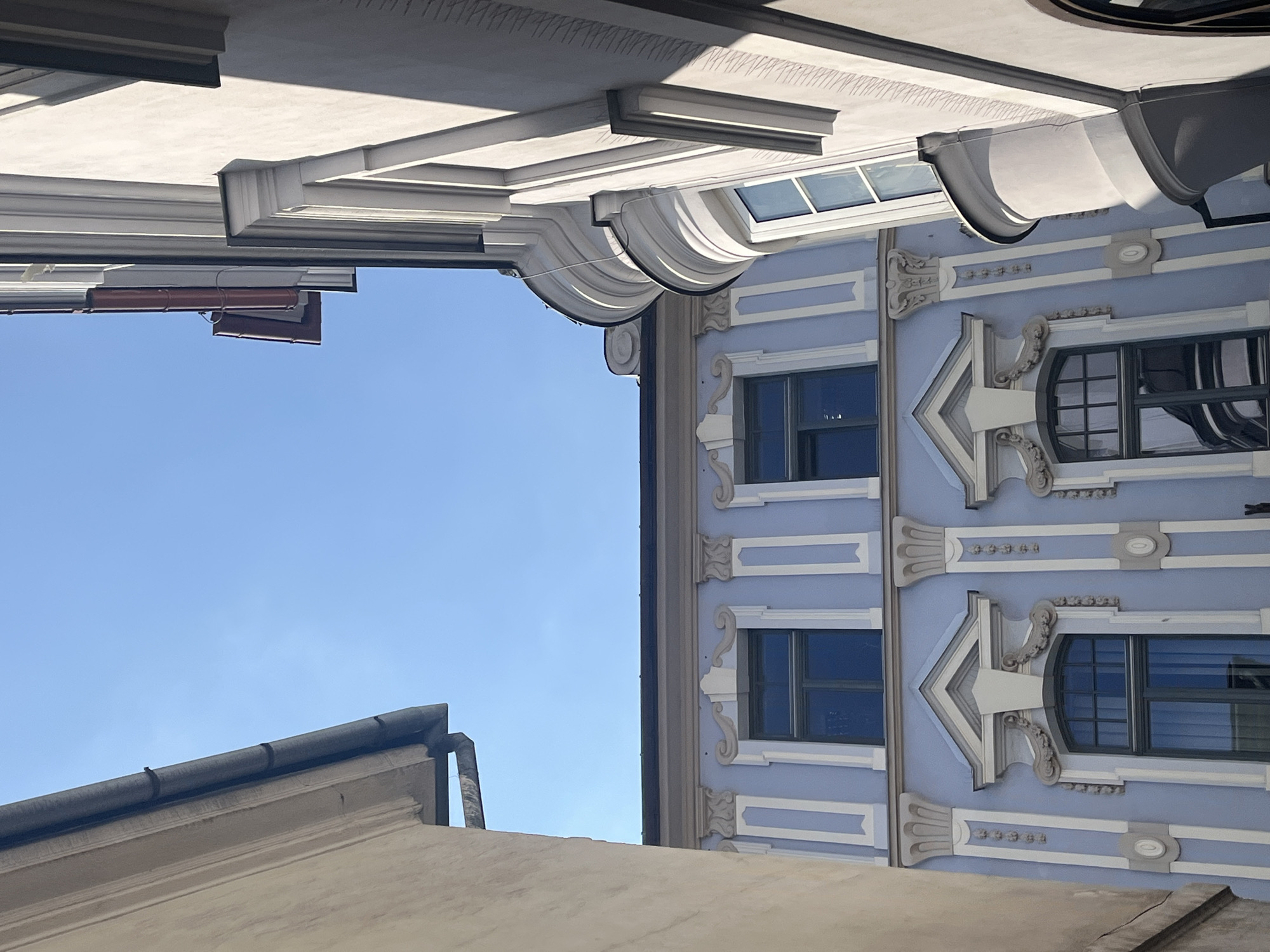 The image shows the exterior of buildings with a clear blue sky. On the left, a cream-colored building with a dark gutter runs along the top. In the center, a light blue building features architectural details around two windows. The upper window is rectangular with a dark frame, and the lower window is arched with a dark frame and decorative elements above it. White vertical trim and decorative columns with ornate capitals frame the windows on the blue building. To the right, a light gray building is angled, with a curved bay window and more white decorative trim. Dark gutters are visible on the roofs of the buildings. The scene appears to be an urban environment, possibly a street or courtyard, given the proximity of the buildings. There are no people or any discernible actions taking place.