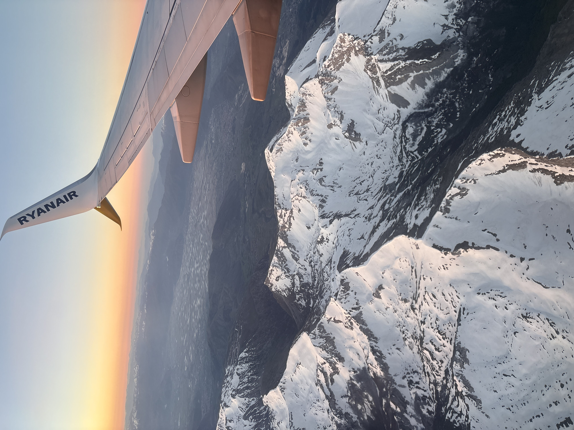 An aerial photograph captures a dramatic view from an airplane window, showcasing the aircraft's wing against a backdrop of snow-capped mountains and a vibrant sky. The image is oriented vertically, with the airplane wing extending from the upper left towards the center. The distinct white "RYANAIR" logo is clearly visible on the wing's surface, indicating a commercial flight.
Below the wing, a vast expanse of rugged, snow-covered mountains dominates the right side of the frame. The peaks and valleys are sharply defined, with patches of dark rock contrasting against the white snow, suggesting deep ravines and steep slopes. Shadows cast by the mountain ridges highlight their dramatic topography, while some areas of snow catch the light, appearing bright and pristine.
On the left, behind the airplane wing, the sky displays a beautiful horizontal gradient of colors, transitioning from warm oranges and yellows at the horizon line to softer blues higher up. This atmospheric effect strongly suggests either a sunrise or sunset, bathing the distant landscape in a soft, ethereal light. The overall impression is one of serene flight over a majestic, wintry mountain range. Given the flight context of Rome, Italy, these mountains are likely part of the Alps or another range within the wider European region. The weather appears clear and calm, offering unobstructed views of the breathtaking scenery below.