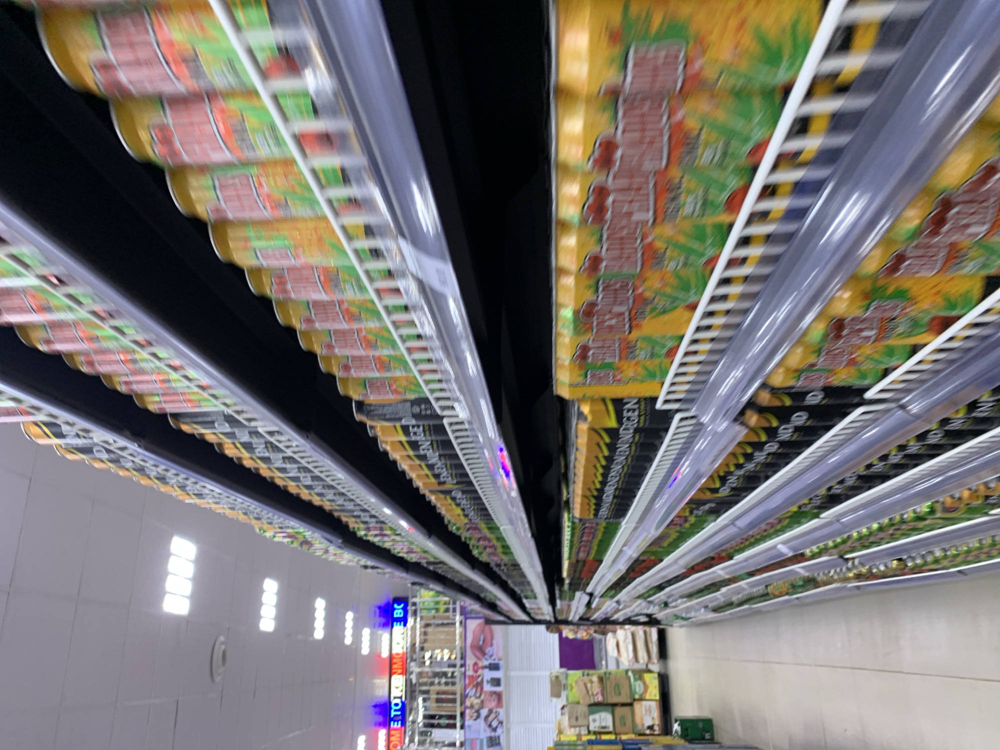 The image depicts a supermarket aisle in Ikorodu, Nigeria. The perspective is from within the aisle, looking towards the back of the store. Rows of shelves are stocked with canned goods, primarily beverages. The shelving units have clear plastic strips along the front. The fluorescent lights are visible in the ceiling. At the end of the aisle are various product displays and shelving units, with a focus on packaged goods and drinks. Some signage is visible in the distance.