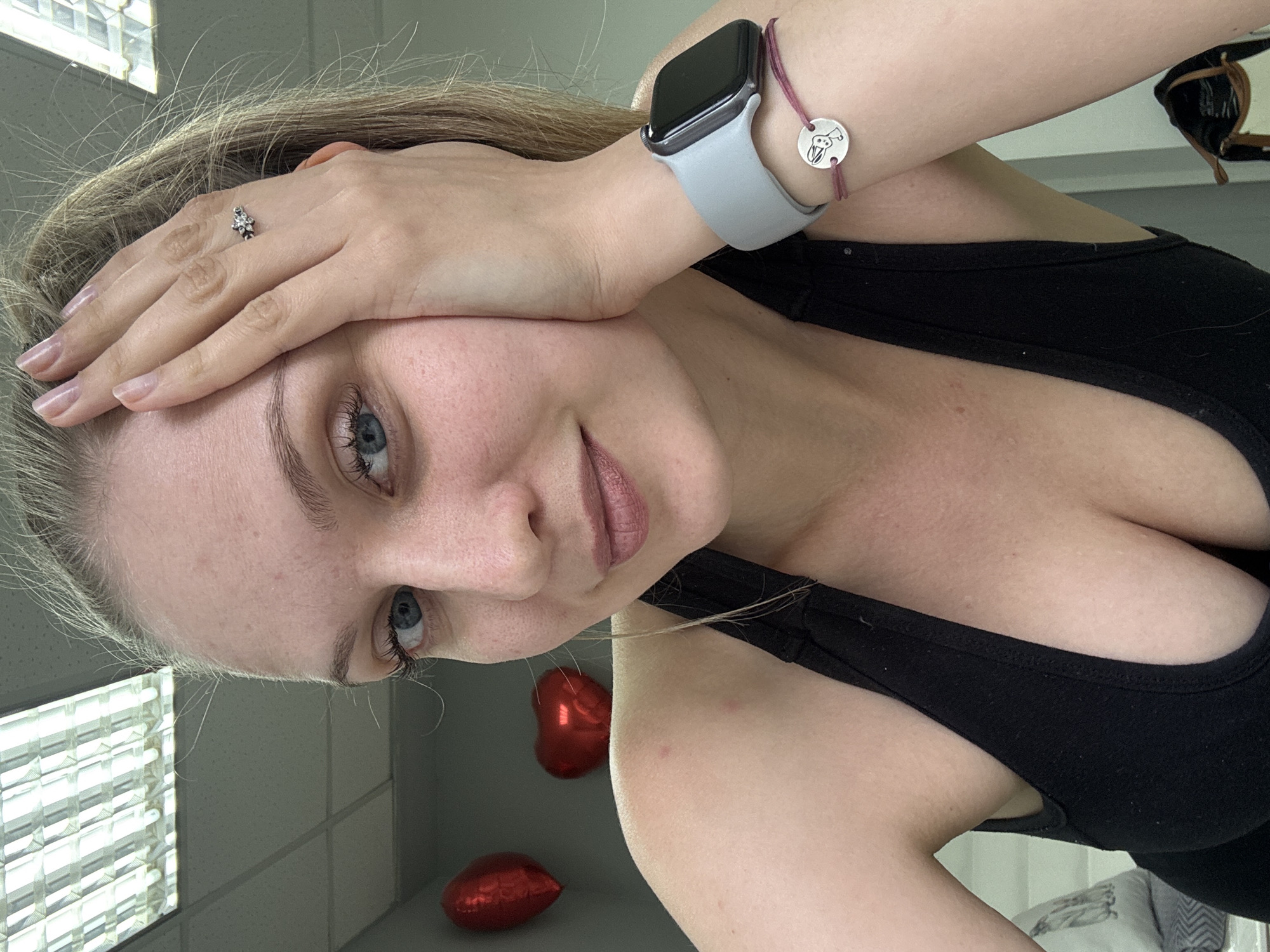 A young woman, appearing to be in her 20s or early 30s, is featured in a close-up, vertically oriented portrait. She has light brown hair, fair skin with some freckles, and blue eyes. Her head is tilted, and her left hand rests gently against her forehead, partially obscuring the top of her face. A soft, subtle smile graces her lips as she gazes towards the camera.
She is dressed in a black tank top or sports bra. On her left wrist, she wears a light grey Apple Watch with a dark screen, alongside a thin red string bracelet adorned with a small, circular silver charm. The charm displays a simplified animal head, resembling a rabbit or a dog. A silver ring with a cluster of small, clear stones is visible on her right ring finger. Her nails are manicured with a natural, light polish.
The indoor setting includes two red, heart-shaped balloons in the background, suggesting a celebratory or personal atmosphere. The ceiling features white tiles and a rectangular fluorescent light fixture, which is also reflected in the upper left of the image. A patterned pillow or blanket is partially visible in the lower right corner. The lighting is bright and direct, emanating from the overhead fixtures. The scene captures a relaxed moment indoors, located in Izhevsk, Russia.
