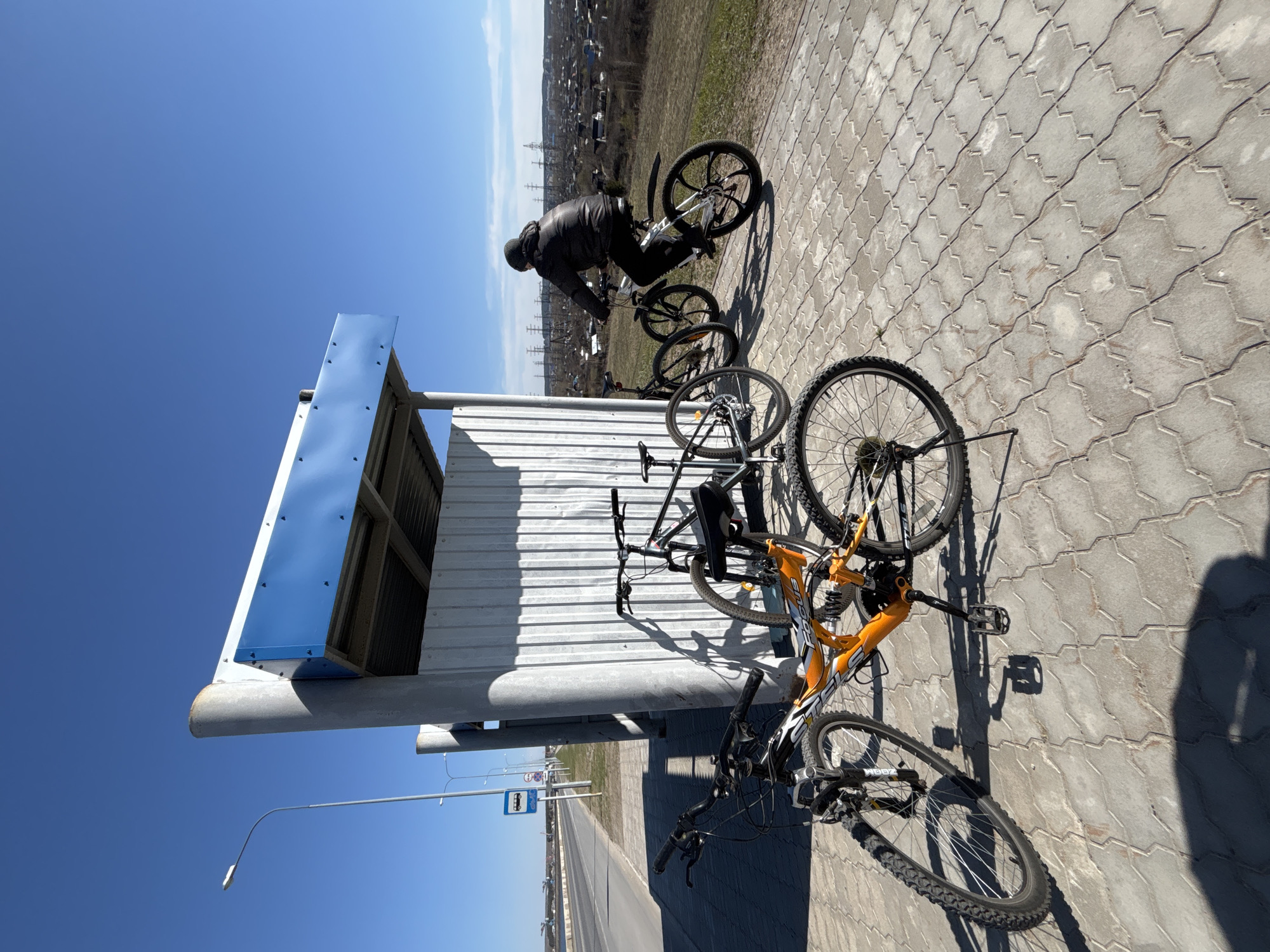 The image shows a street scene in what appears to be Chaikovsky, Russia, based on the bus stop sign and general architecture. The view is from a low angle and is slightly tilted. The foreground is dominated by a bus stop with a blue and white roof. There are three bicycles parked on the paved area next to the bus stop. One bicycle is predominantly orange. In the background, a person wearing dark clothing is riding a white bicycle on the sidewalk. In the distance, there is a cityscape with buildings, trees, and utility poles under a clear blue sky. The pavement is made of interlocking concrete blocks.