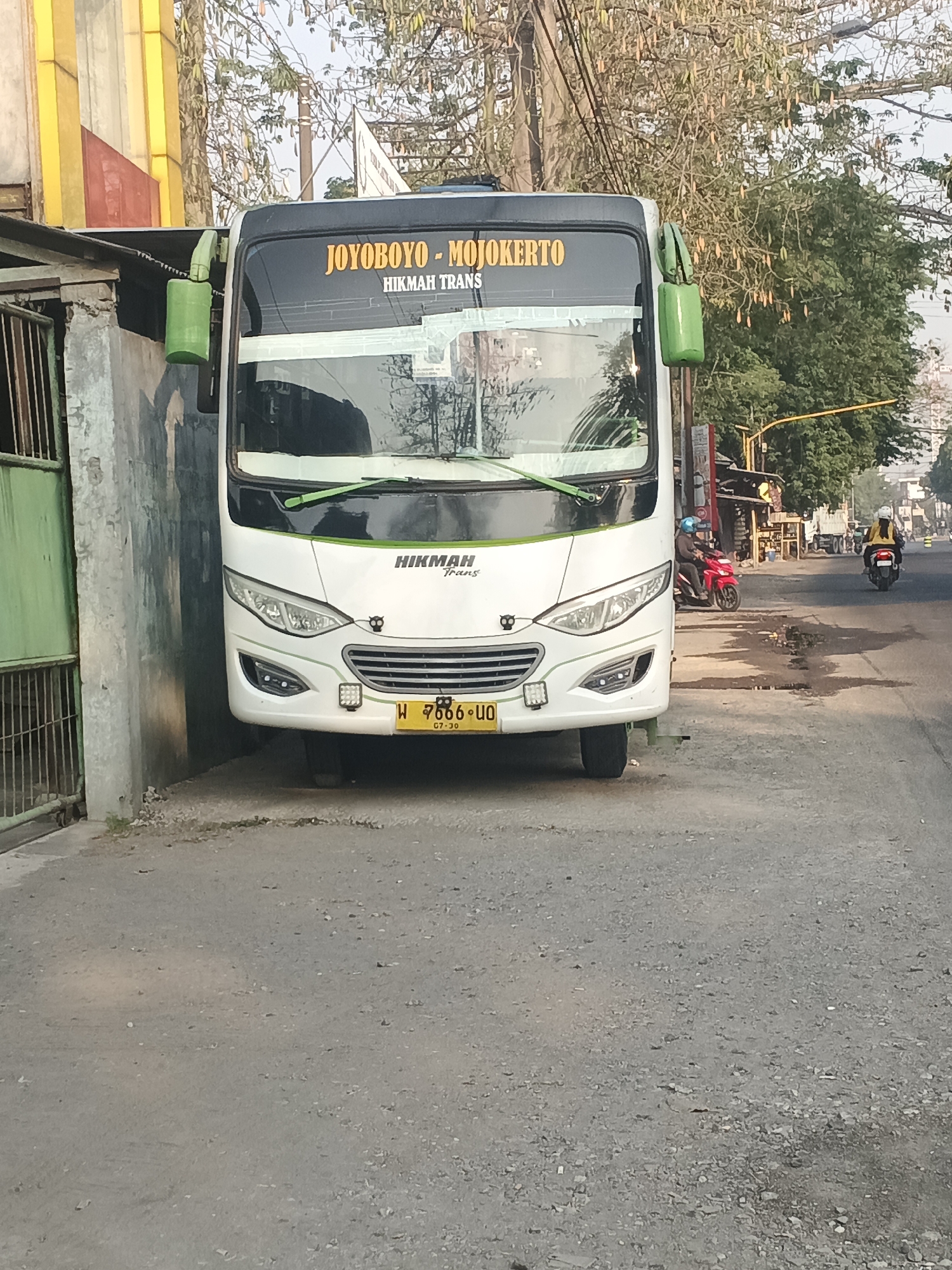 A white bus with a black and green trim is parked on the side of a paved road. The bus has "JOYBOYO - MOJOKERTO" and "HIKMAH TRANS" written on the windshield in yellow and black lettering. A license plate on the front of the bus reads "W 7666 UO". The bus is partially obscured by a grey concrete wall on its left. A green metal gate is visible in front of the wall. Trees with dry branches and seed pods are in the background. The road is littered with debris and puddles of water. Two motorcycles are on the road, one red and one black, each with a rider. The rider of the red motorcycle is wearing a blue helmet, and the rider of the black motorcycle is wearing a yellow jacket and a white helmet. In the distance, a white truck is visible. The scene appears to be in a rural or semi-urban area in Kemangsen, Indonesia, likely during daylight hours given the lighting conditions.