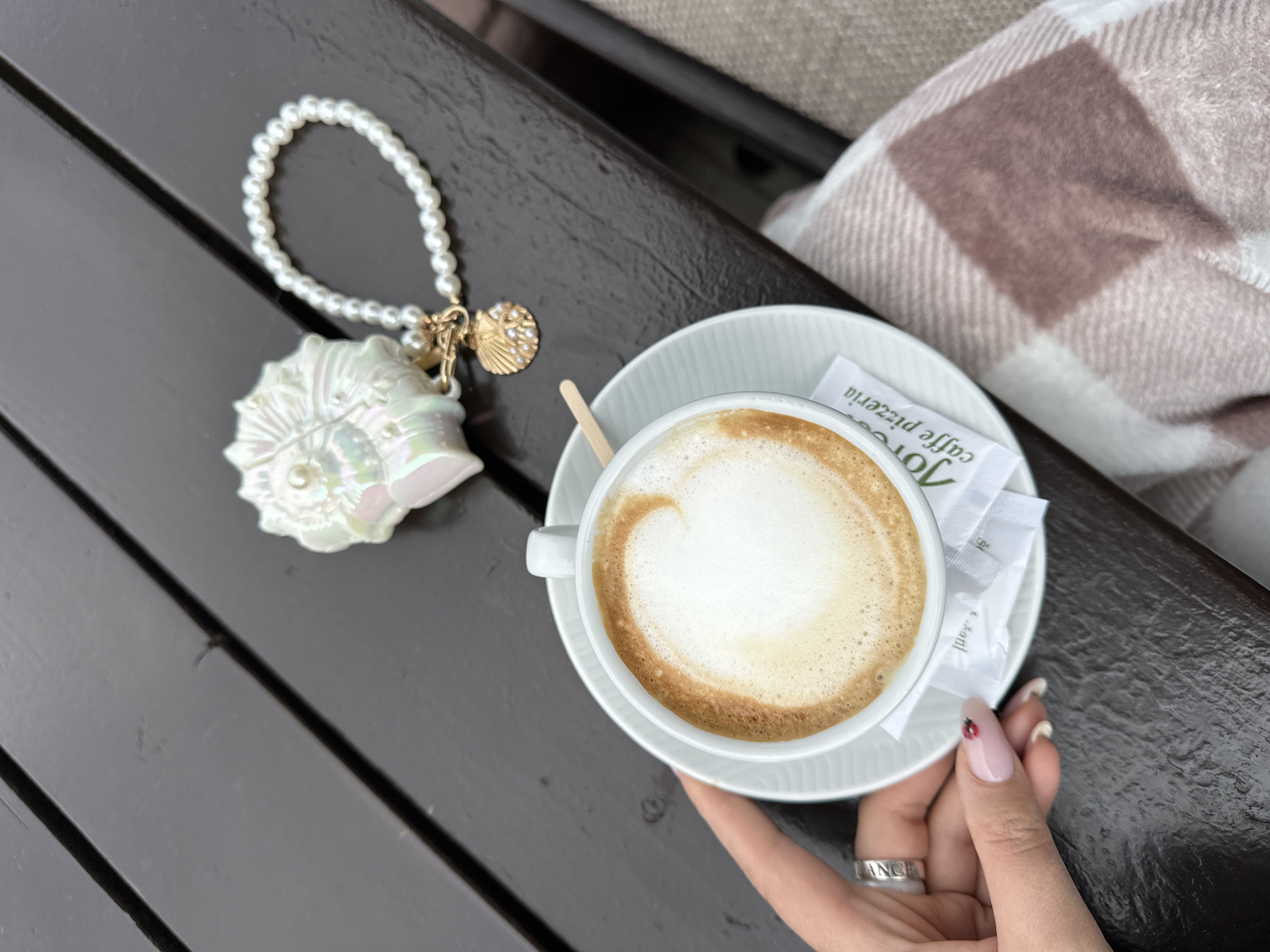 A high-angle shot shows a hand holding a cup of coffee on a saucer placed on a dark brown wooden surface, likely a bench. The coffee has a light brown foam top. Next to the cup is a small, white paper packet. To the left of the cup is a bracelet composed of faux pearls with an iridescent seashell and gold seashell charm. A tan and white plaid blanket is partially visible in the upper right corner. The hand holding the cup has manicured nails and wears a silver ring with the word "ANGEL" inscribed on it. The image was taken in Šušanj, Montenegro.