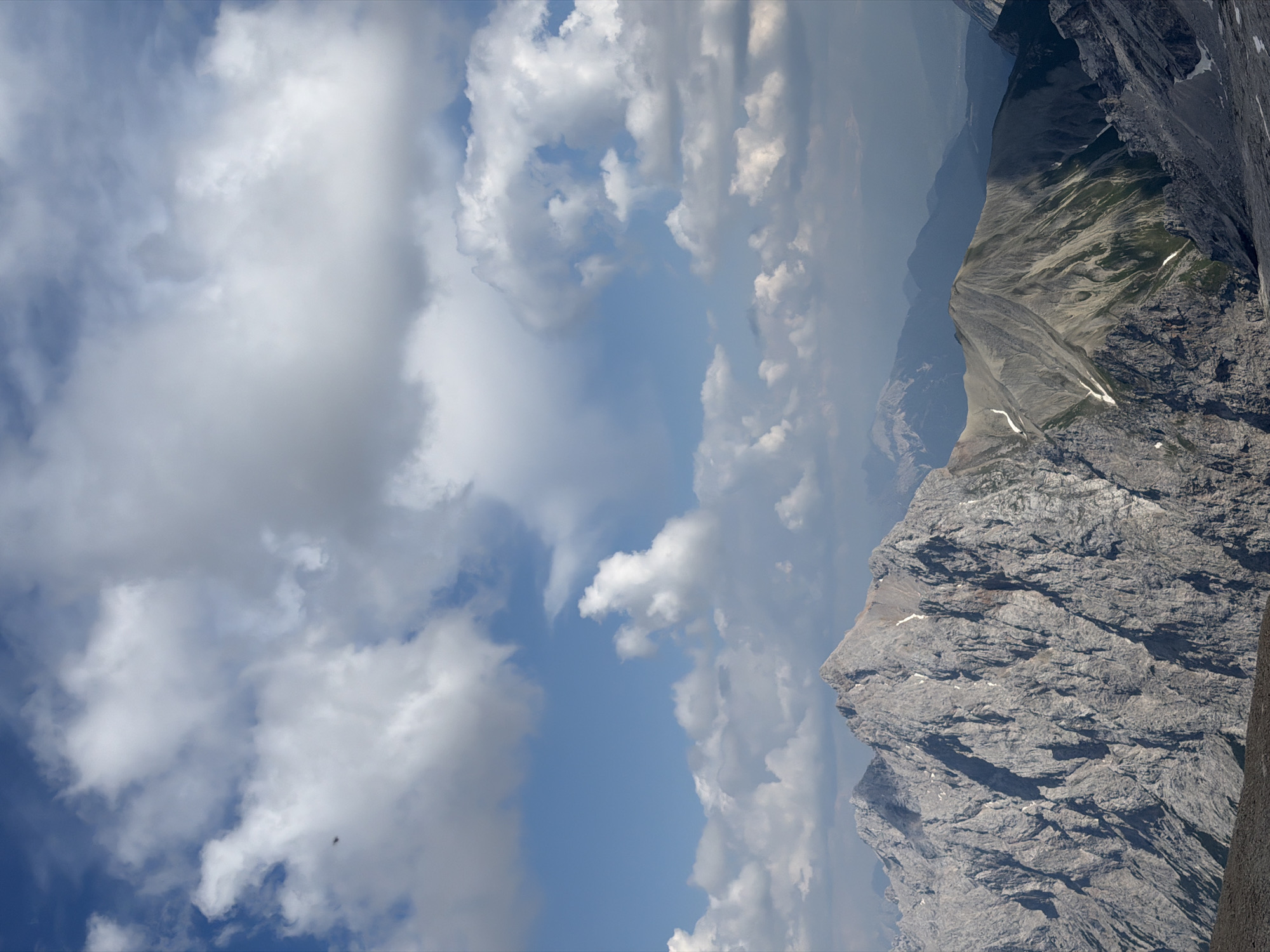 The image depicts a majestic mountainous landscape with a vast sky filled with white, fluffy clouds. The foreground is dominated by rugged, grey mountain peaks, sharply defined and showing signs of weathering. Some patches of green vegetation are visible on the slopes of the middle ground mountains, interspersed with areas of snow or lighter-colored rock. In the distance, layers of progressively hazier mountains fade into the horizon under the expansive sky. The scene is likely a high-altitude vista, characteristic of alpine regions. Based on the description of Garmisch-Partenkirchen, Germany, this image could be a view of the Bavarian Alps, possibly including peaks like the Zugspitze.