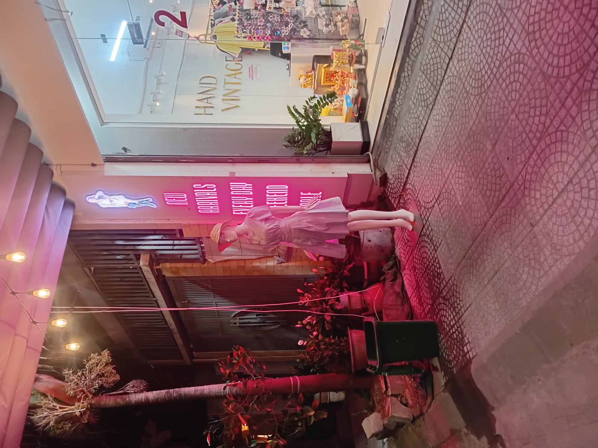 A mannequin wearing a light purple dress and a straw hat stands on a sidewalk in front of a shop. The shop has a neon sign that says "NEW ARRIVALS EVERY DAY WEEKEND SALE". Inside the shop, there are clothes racks and shelves displaying various items. A large red number "2" is visible on the shop window. The sidewalk is paved with patterned tiles and is illuminated by pink neon lights. There are plants and a green trash can on the sidewalk. The scene appears to be a street in the city of Đà Nẵng, Vietnam, at night.