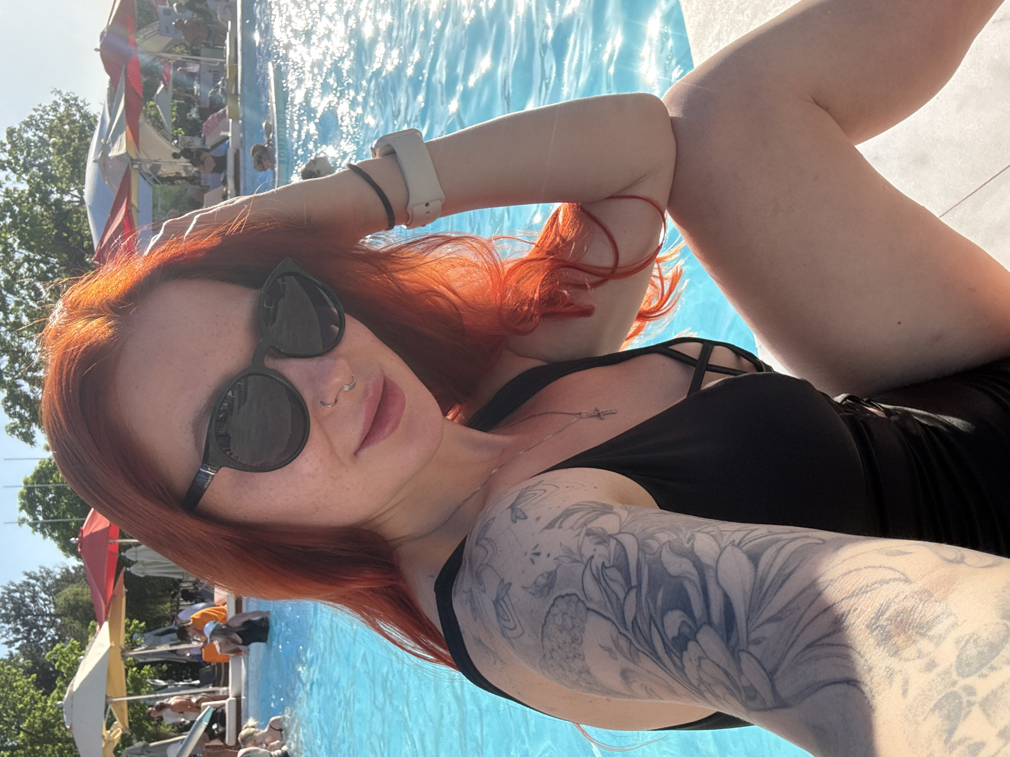 A red-haired woman is lying by a pool in Rostov-on-Don, Russia. She is wearing a black swimsuit and sunglasses. She has a tattoo on her left arm. The pool is surrounded by people and trees.