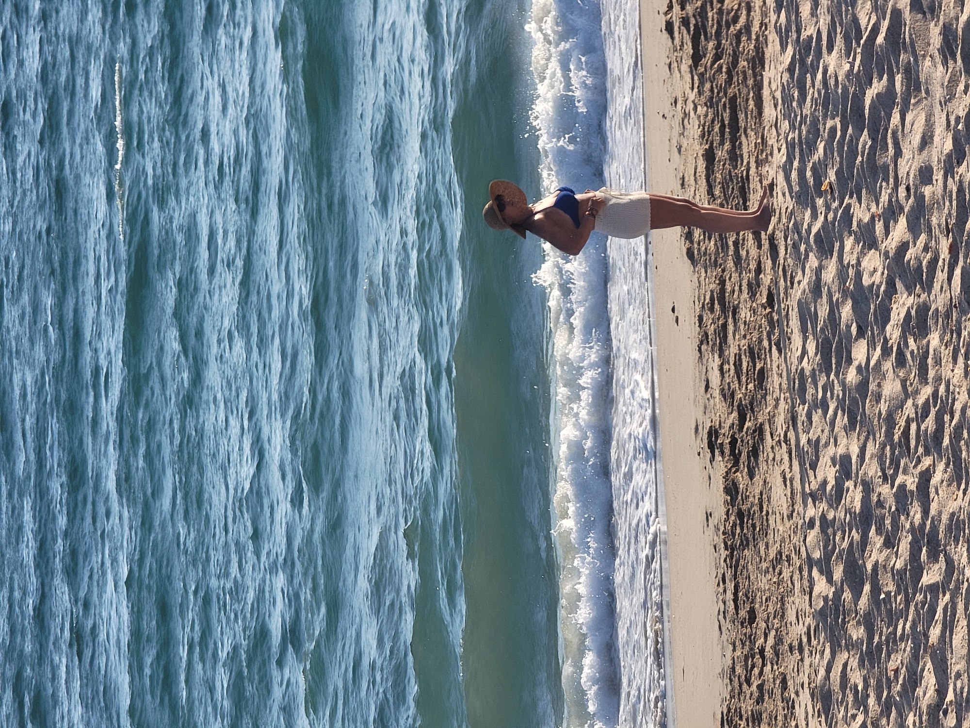 A woman stands on a sandy beach, facing the ocean. She is wearing a blue bikini top and white shorts. A straw hat with a wide brim shades her face. The ocean behind her is choppy with white waves breaking near the shore. The sand is textured with ripples and shadows. The scene appears to be located on a beach in Quarteira, Portugal.
