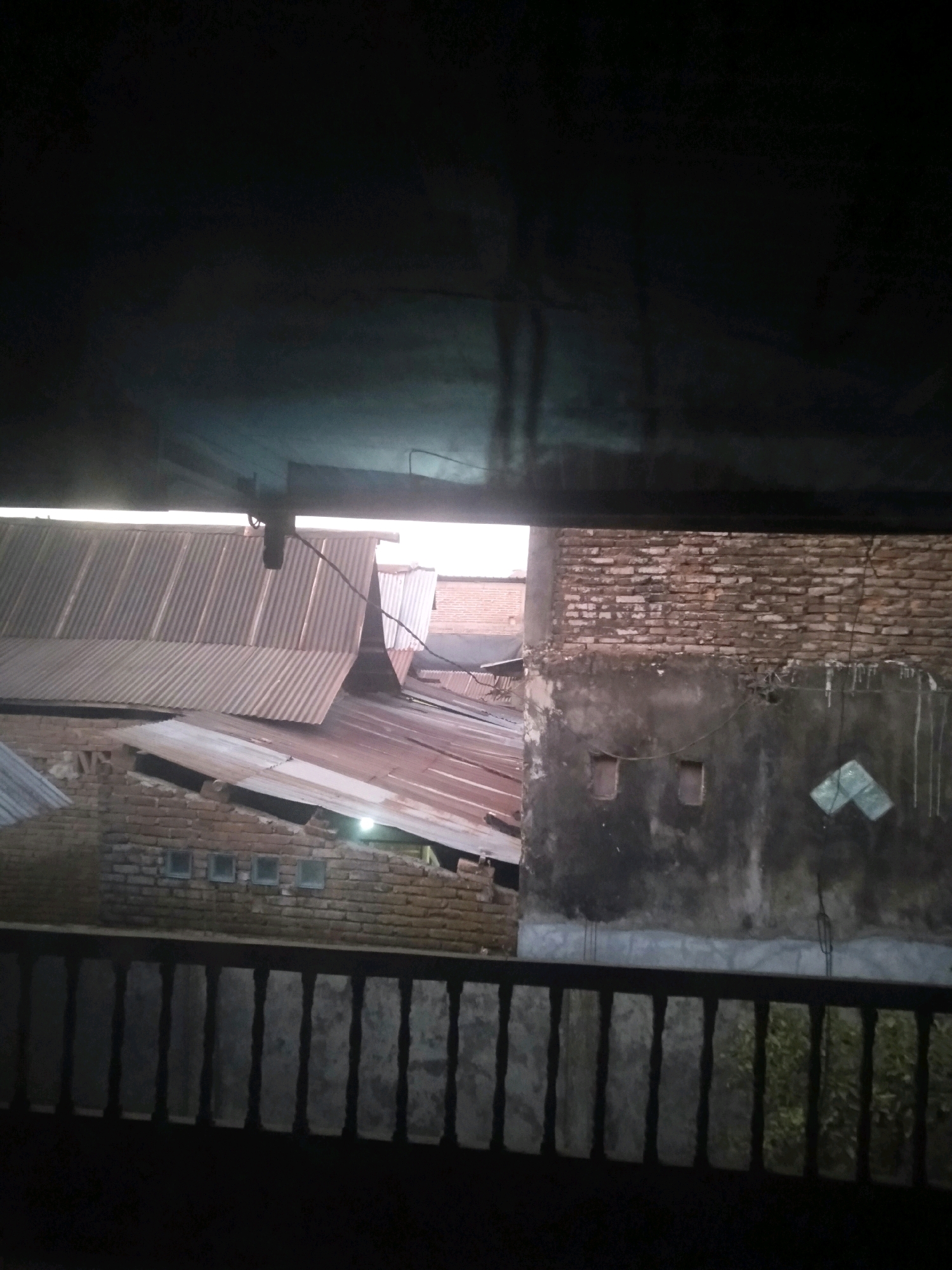 This image depicts a residential area in Pallangga, Indonesia, at dusk or night. The foreground features a dark, blurry railing, suggesting the vantage point is from a porch or balcony. Beyond the railing, corrugated metal roofs of several houses are visible, some appearing weathered and discolored. The walls of the buildings are constructed from brick, with some sections covered in a dark, textured plaster. Small, square windows are present in some of the walls. A green object, possibly a plastic bag or a piece of cloth, is attached to the wall on the right. The overall lighting is dim, with some artificial light sources casting a faint glow. The scene appears to be a typical urban or semi-urban residential environment in Indonesia.