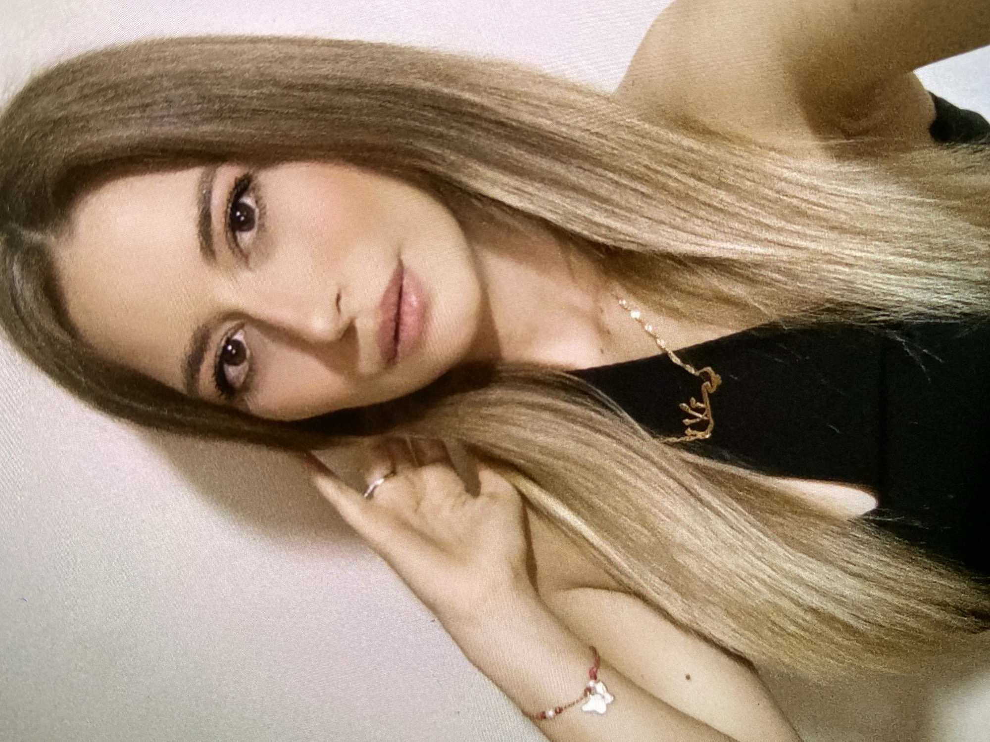 The media file presents a portrait of a young woman with long, straight, light brown and blonde hair. Her head is tilted, and she gazes directly at the camera with a soft, gentle expression. She has dark brown eyes and well-defined eyebrows, complemented by subtle, natural-looking makeup on her eyes and lips.
She is dressed in a black, sleeveless top. Around her neck, she wears a delicate gold-colored necklace featuring a script pendant that appears to spell out a name, possibly "Zara." On her left hand, a simple silver ring is visible on one of her fingers. Her left wrist is adorned with a red string bracelet, from which hangs a small, white, butterfly-shaped charm. Her left arm is bent, with her hand positioned near her hair and face.
The background is a plain, light-colored surface, suggesting an indoor setting. The lighting is soft and even, highlighting her features without harsh shadows. The image captures a serene moment, possibly a casual self-portrait or a posed photograph. There are no overt location cues within the image itself to confirm it is Bankya, Bulgaria, but this is the provided context. No specific activity other than posing is depicted, and there are no indicators of the time of day or weather. The only visible text is on the necklace pendant.