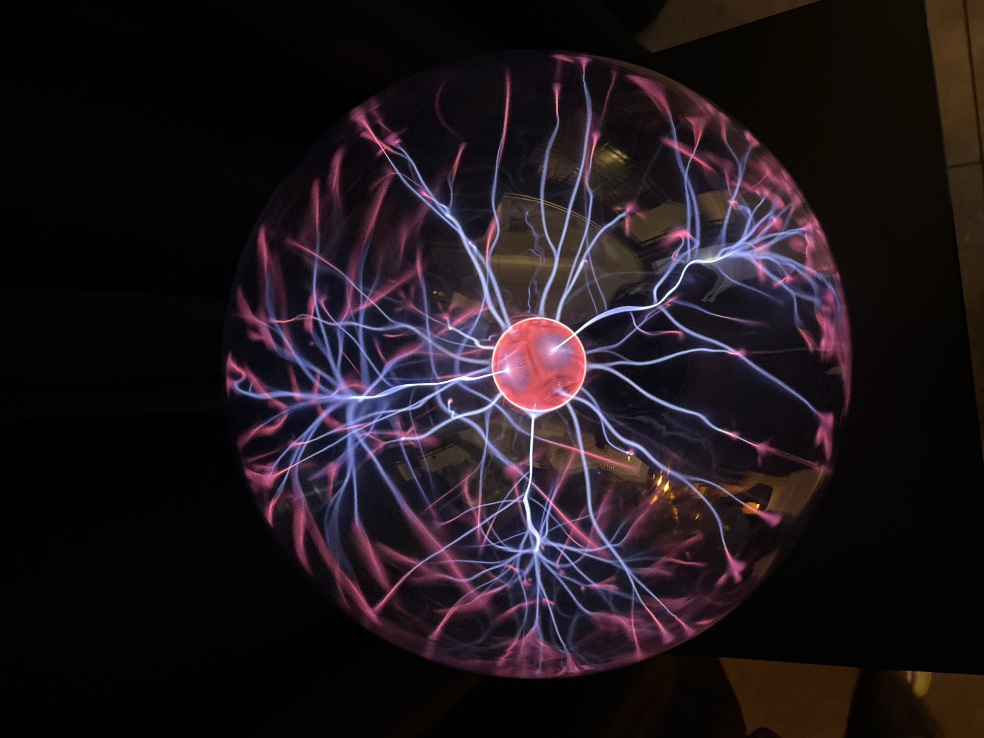 The image depicts a plasma ball on a dark surface. The plasma ball has a red sphere at its center, with tendrils of purple and pink light emanating outward. The background is predominantly black, suggesting a low-light environment. There is no information indicating the location is Unknown, Unknown or any other location. No people are visible in the image.