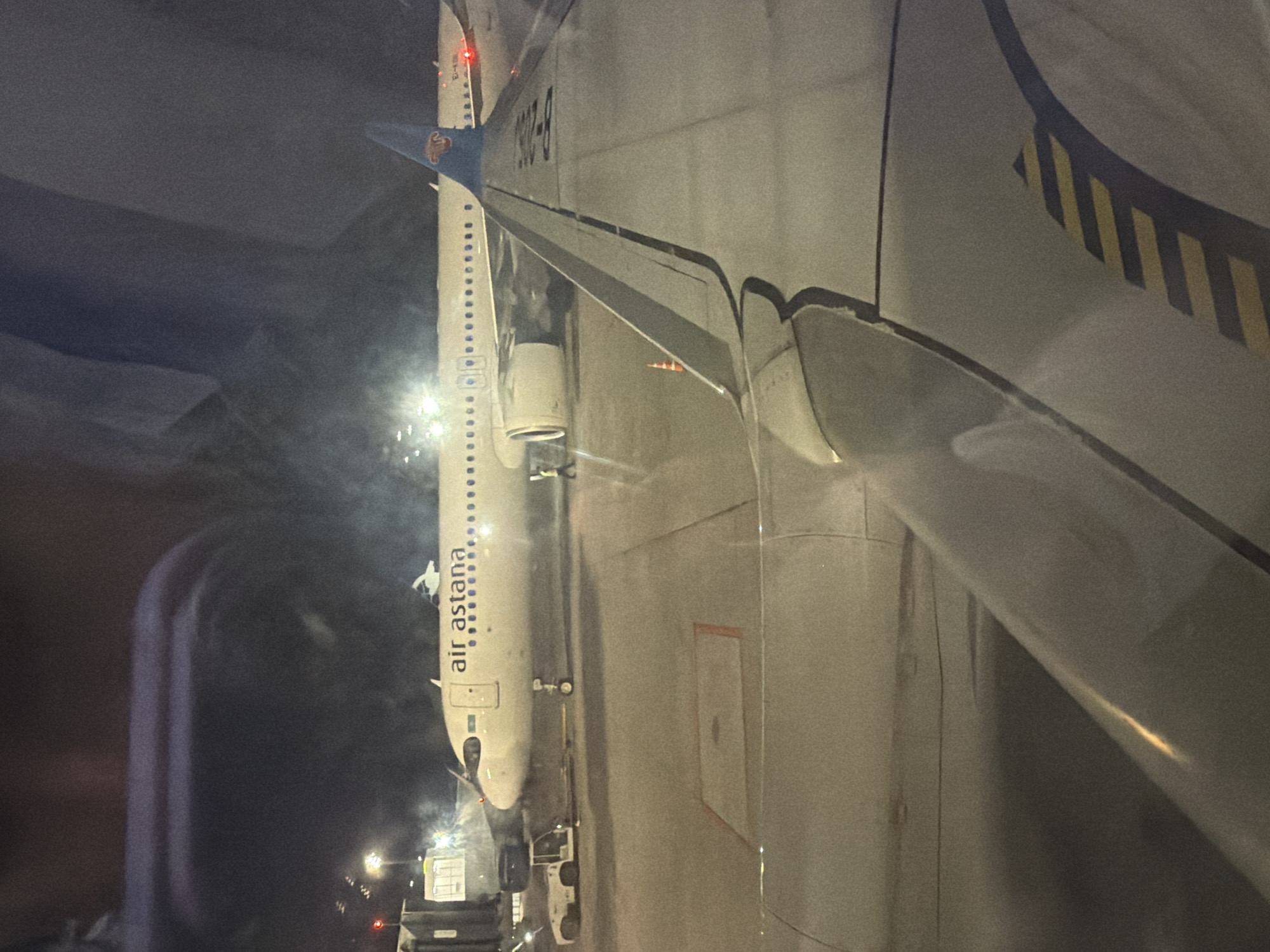 The image depicts an Air Astana aircraft at night. The aircraft is positioned near what appears to be ground support vehicles. The location is inferred to be Almaty, Kazakhstan based on the airline. The aircraft features a light-colored fuselage with the "Air Astana" logo and the tail fin displaying the airline's branding. Part of the wing and fuselage of another aircraft, with yellow and black markings, is also visible in the foreground. The environment appears to be dimly lit, possibly at an airport apron or gate.