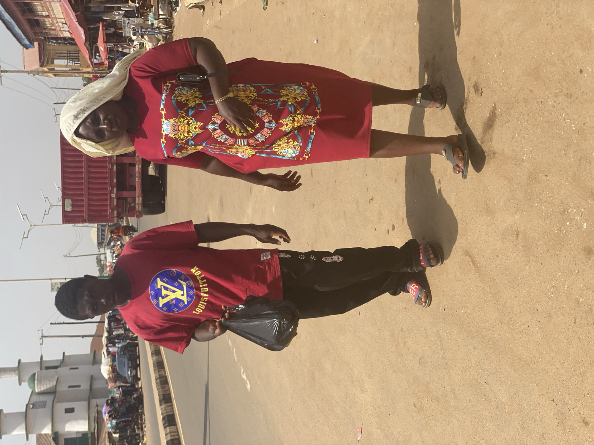 The image depicts two people standing on a paved surface. A woman in a red dress with a gold and blue design, and a white head covering, is on the upper part of the image. She is wearing sandals and has a small black purse on her arm. A man in a red Louis Vuitton t-shirt and black pants is standing below her. He is carrying a black plastic bag and is also wearing sandals. In the background, there are buildings, market stalls, and vehicles suggesting an urban environment. There are structures of the Kafanchan B, Nigeria city.