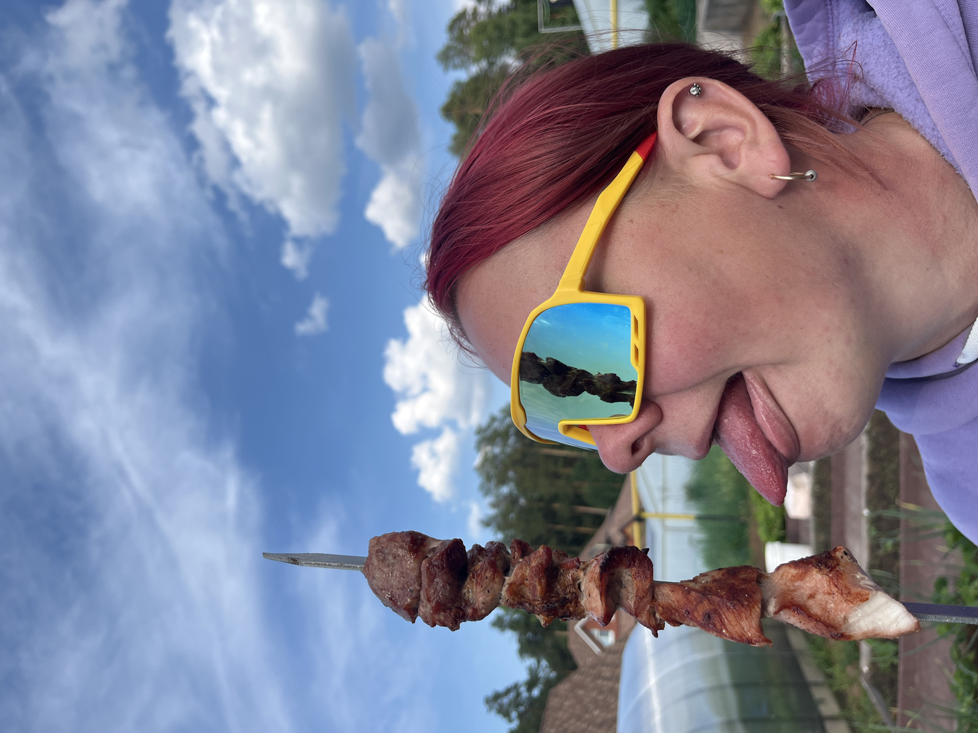 A woman with red hair and yellow sunglasses sticks out her tongue towards a skewer of grilled meat. She is wearing a purple hoodie and has earrings in both ears. The background shows a garden with a greenhouse and trees under a cloudy blue sky. The location is not explicitly provided in the image or crops.