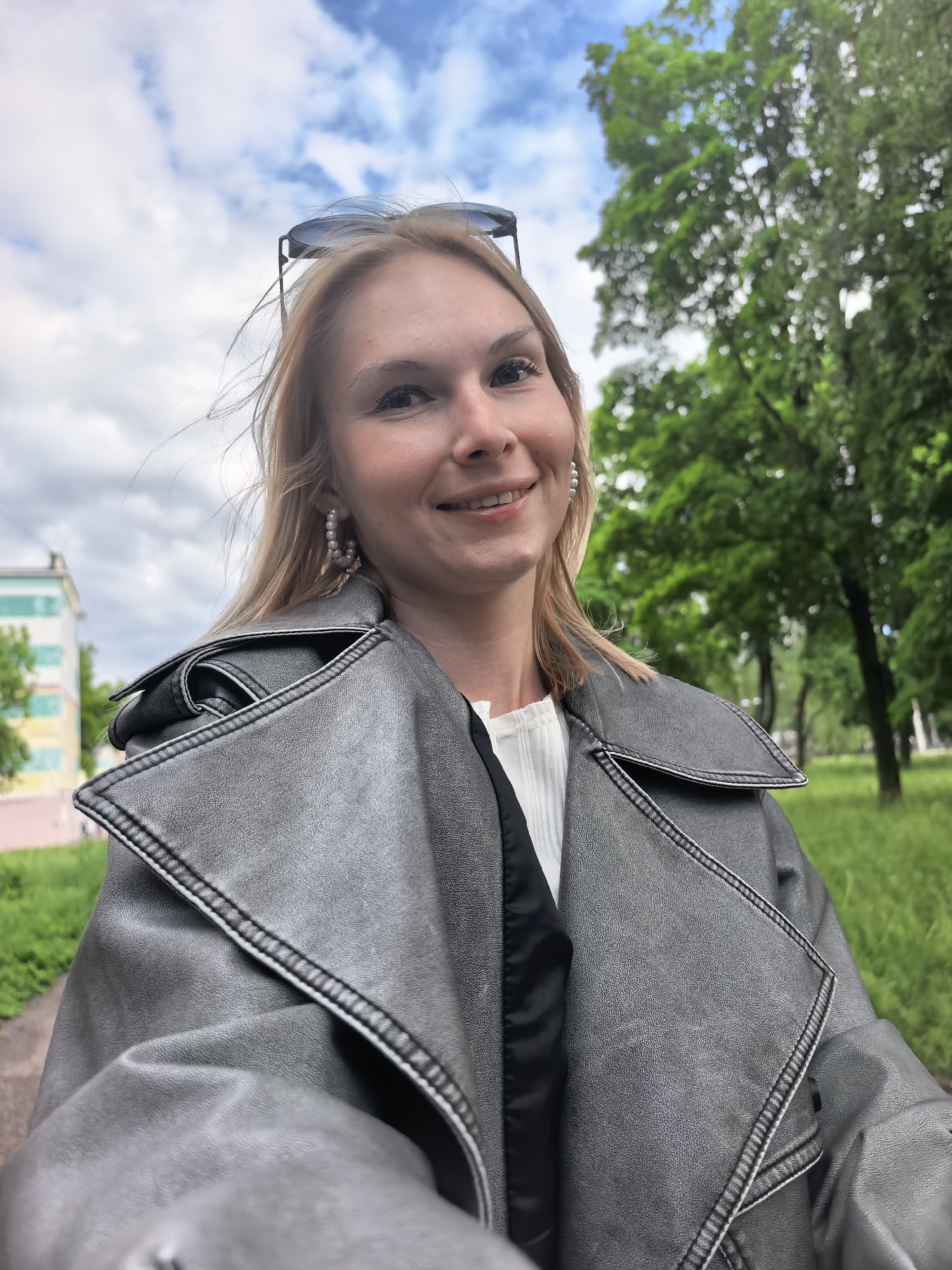 The image shows a woman in Sterlitamak, Russia, wearing a grey leather trench coat and pearl earrings. She has blonde hair and sunglasses on her head. The background includes a cloudy sky, green trees, and a building with green and yellow trim. The photo appears to be taken outdoors in a park or green space within the city.