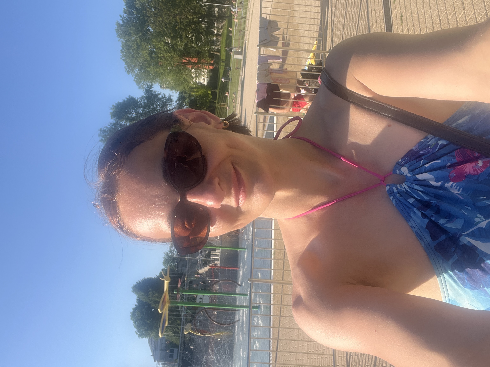 A woman, wearing sunglasses and a blue and pink patterned bikini, takes a selfie outdoors on a sunny day. She has her hair pulled back and is smiling. Behind her, a shallow water park with spray features is visible, as well as a fence and several people relaxing in the background. The location is a public park on a clear day.