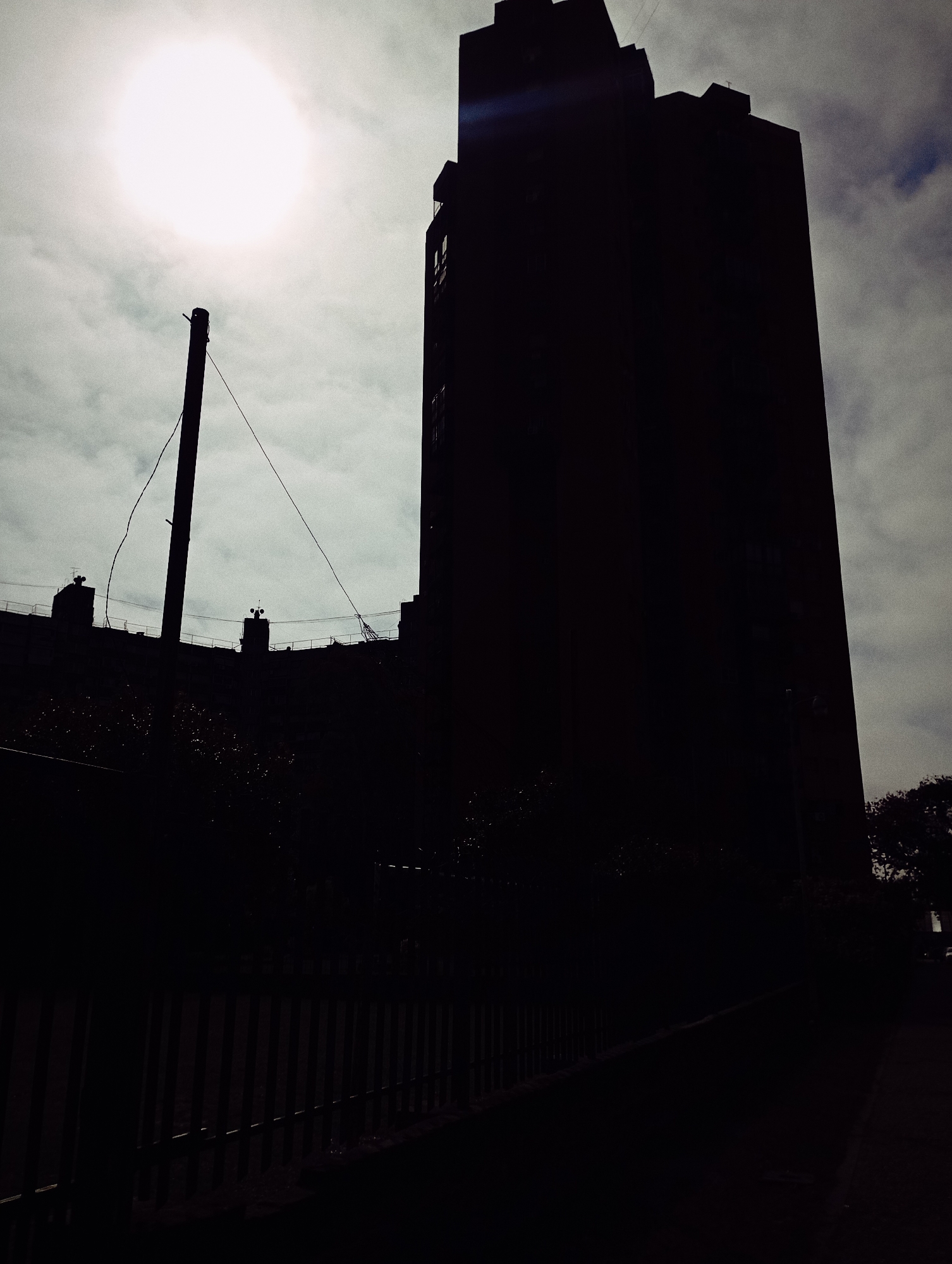 The image shows a silhouette of a tall building against a bright, overcast sky. A utility pole with wires is also silhouetted on the left. In the foreground, there is a fence and some foliage. The location is likely an urban area, but the exact location within Buenos Aires, Argentina, cannot be determined from this image. The image contains no people. The subjects include a building, utility pole, and fence. The action is static, depicting a scene rather than an event. There are no observable interactions between subjects. The environment is overcast and urban.