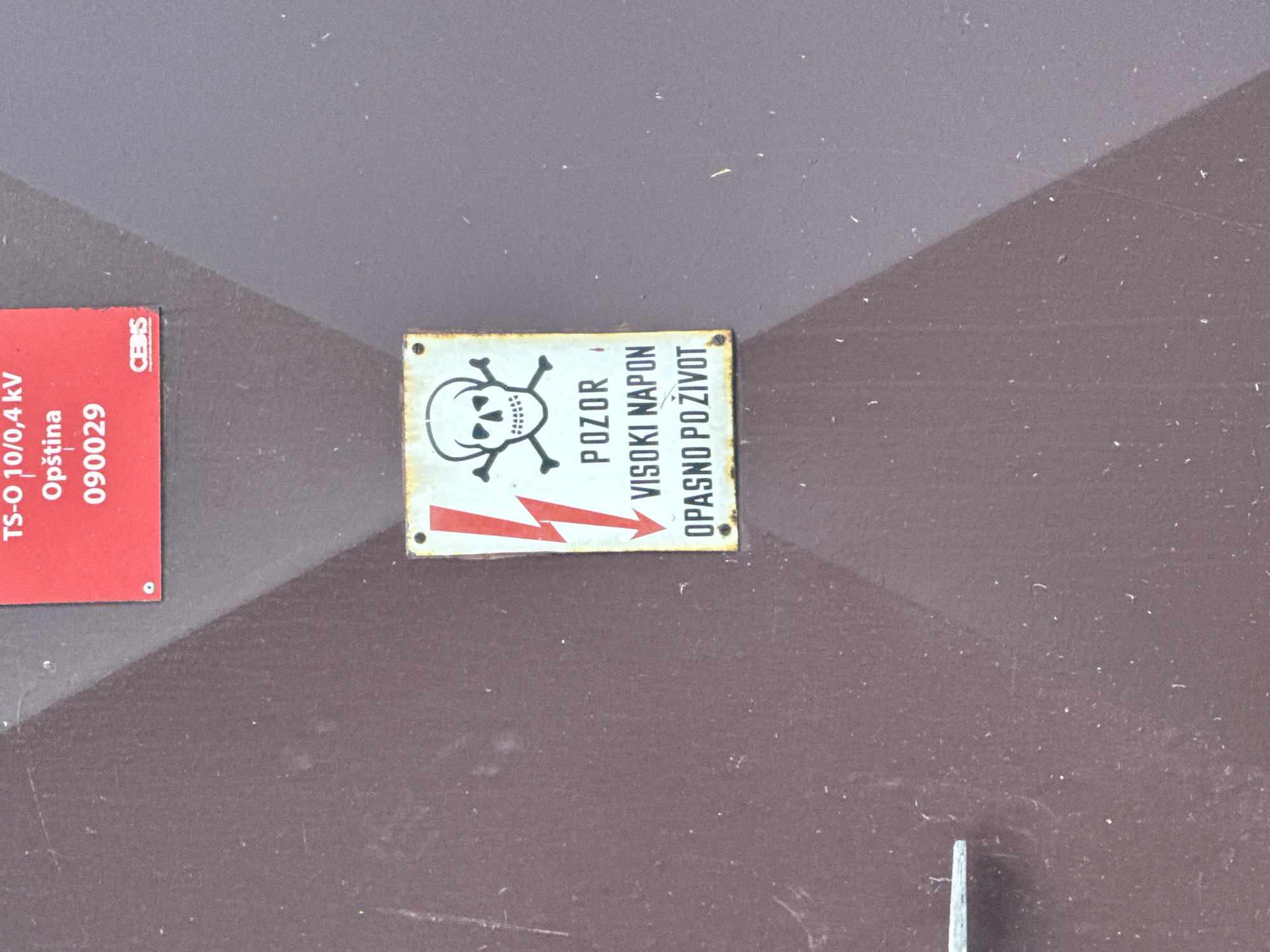 The image shows a close-up of a brown, textured metal door. A red and white warning sign is affixed to the door. The sign displays a skull and crossbones symbol, a lightning bolt, and text in a dark font. The text on the sign reads "POZOR VISOKI NAPON OPASNO PO ŽIVOT", which translates to "CAUTION HIGH VOLTAGE DANGEROUS TO LIFE". Above this sign, a portion of another red sign is visible with white text that reads "TS-O 10/0,4 kV Opština 090029". A small "CEHMS" logo is also visible on this red sign. The door has a metal handle on the left side. The overall environment appears to be outdoors, likely on a utility box or electrical cabinet. The specific location in Budva, Montenegro is not explicitly determinable from the image alone.