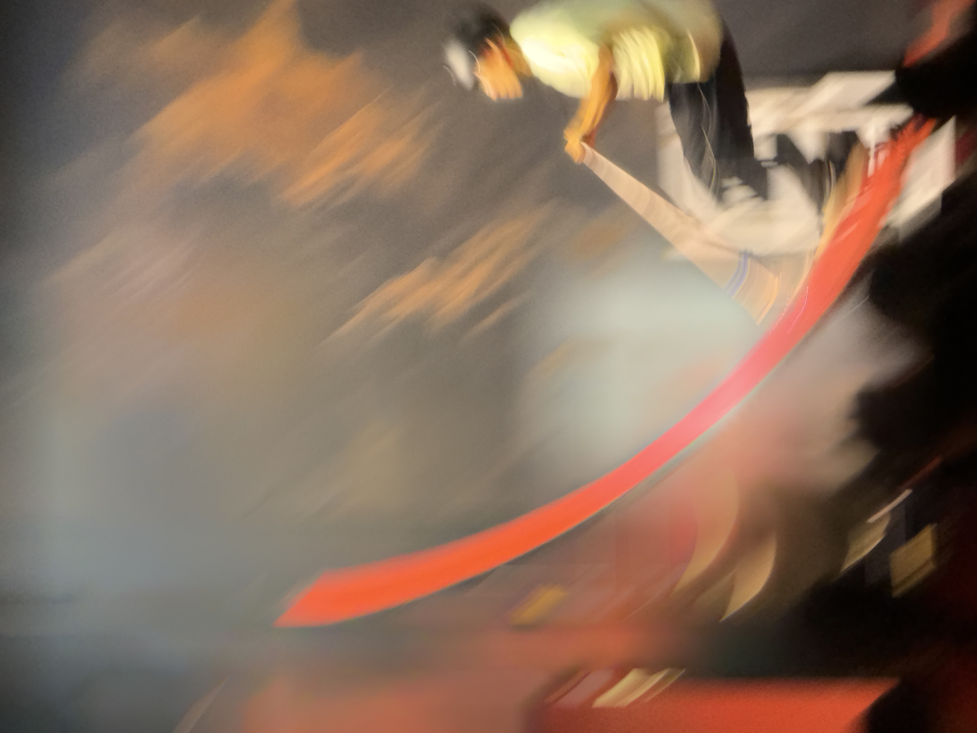 The image shows an abstract, motion-blurred scene. A person in a yellow shirt and dark pants is visible on the right, seemingly in mid-air, holding onto a wooden beam. A vibrant red, curved object, possibly a fabric or ribbon, dominates the left side of the frame. The background is a blur of warm oranges, browns, and grays, suggesting dramatic lighting or stage effects. The overall impression is dynamic and energetic, possibly capturing a performance or acrobatic act. There is no discernible information in the image to indicate the location as Manavgat, Turkey.