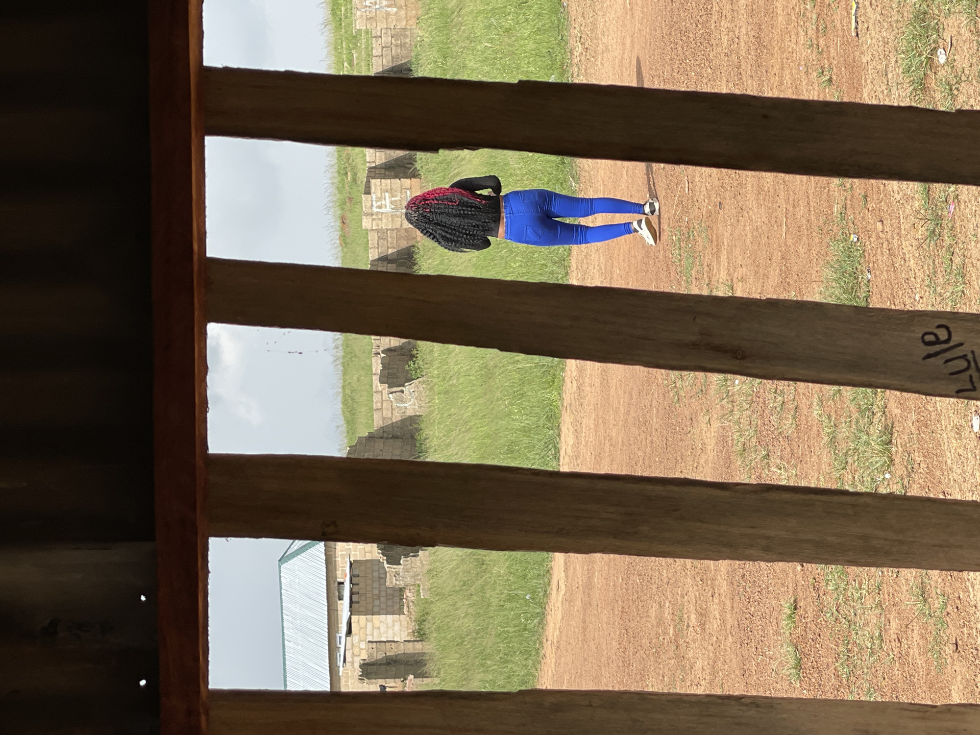 The image shows a person standing in a dirt area with grass and some buildings in the background. The person is viewed from behind and is wearing a black top and bright blue leggings. They have long, dark braided hair with some red highlights. The scene appears to be outdoors, possibly in a rural or semi-urban setting in Durumi, Nigeria. The lighting suggests it is daytime. There is some writing on a wooden beam in the foreground, but it is mostly illegible.