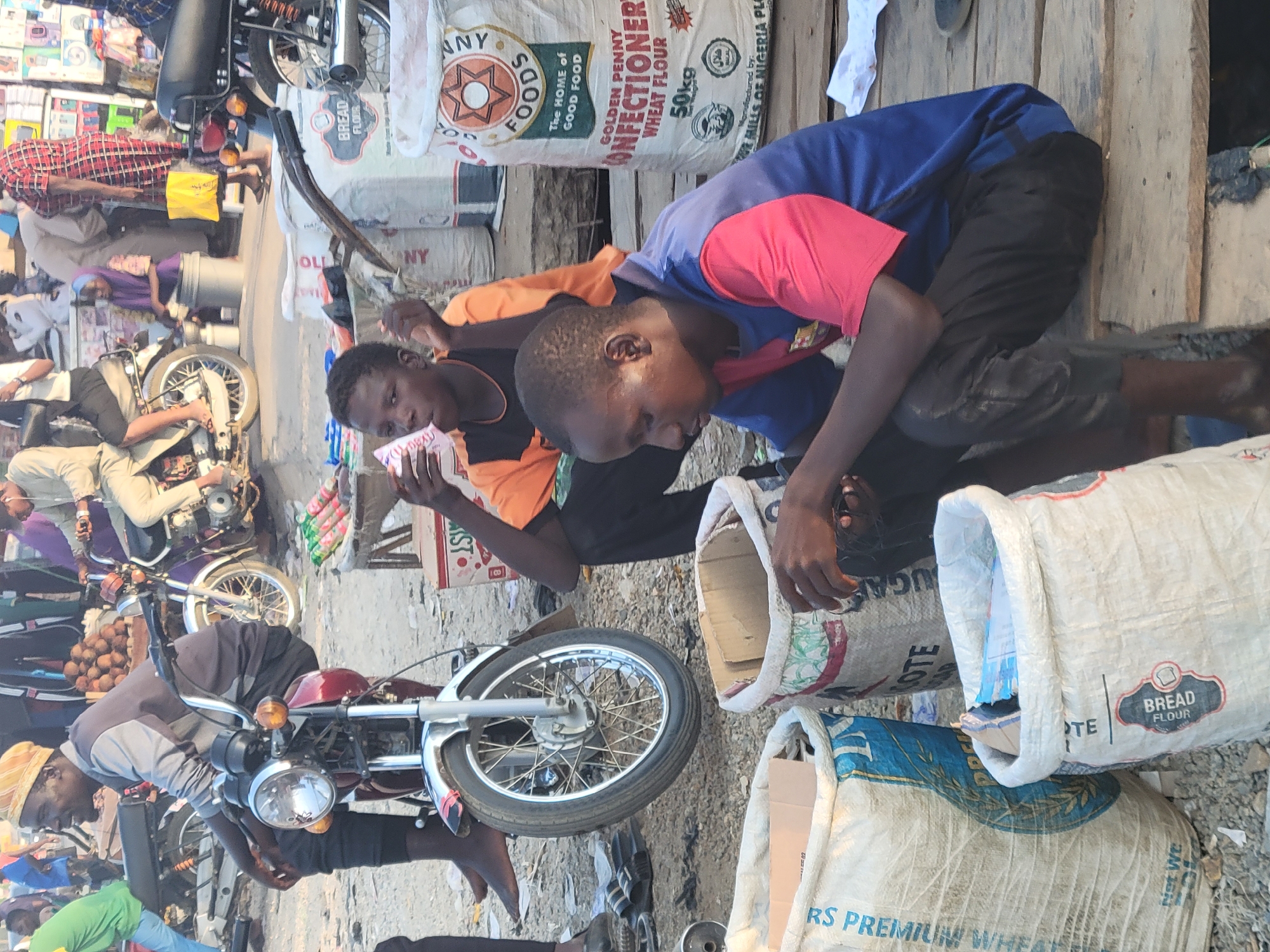 This media file captures a bustling outdoor market scene in Madalla, Nigeria, during bright daylight hours. The image is oriented vertically, depicting various people, goods, and motorcycles.
In the foreground, two young males are prominent. One, wearing an orange and black shirt, holds a stack of cash. The other, dressed in a blue and red shirt, bends over, looking into an open, large sack made of woven material, possibly retrieving or placing items. To their left, a red and silver motorcycle is parked, with a person in a grey top and yellow head covering appearing to be working on it. Another individual, partially obscured, stands next to the motorcycle.
The market setting is dynamic, with multiple other people in the mid-ground and background. One person in a red plaid shirt and yellow bag is seen moving past another parked motorcycle with a purple wheel. Goods are stacked, including sacks, boxes, and a pile of round, brown fruits resembling coconuts.
Several large sacks are visible, some standing upright, others lying down. Visible text on these sacks includes "GOLDEN PENNY CONFECTIONERY WHEAT FLOUR 50kg," "ANY FOODS The HOME of GOOD FOOD," and "BREAD FLOUR." One sack also mentions "Manufactured by: FUGE HILLS OF NIGERIA PLC" and displays a Halal symbol. The ground is a mix of dirt and scattered debris, typical of an informal market. The overall impression is one of commerce and daily activity in an open-air environment.