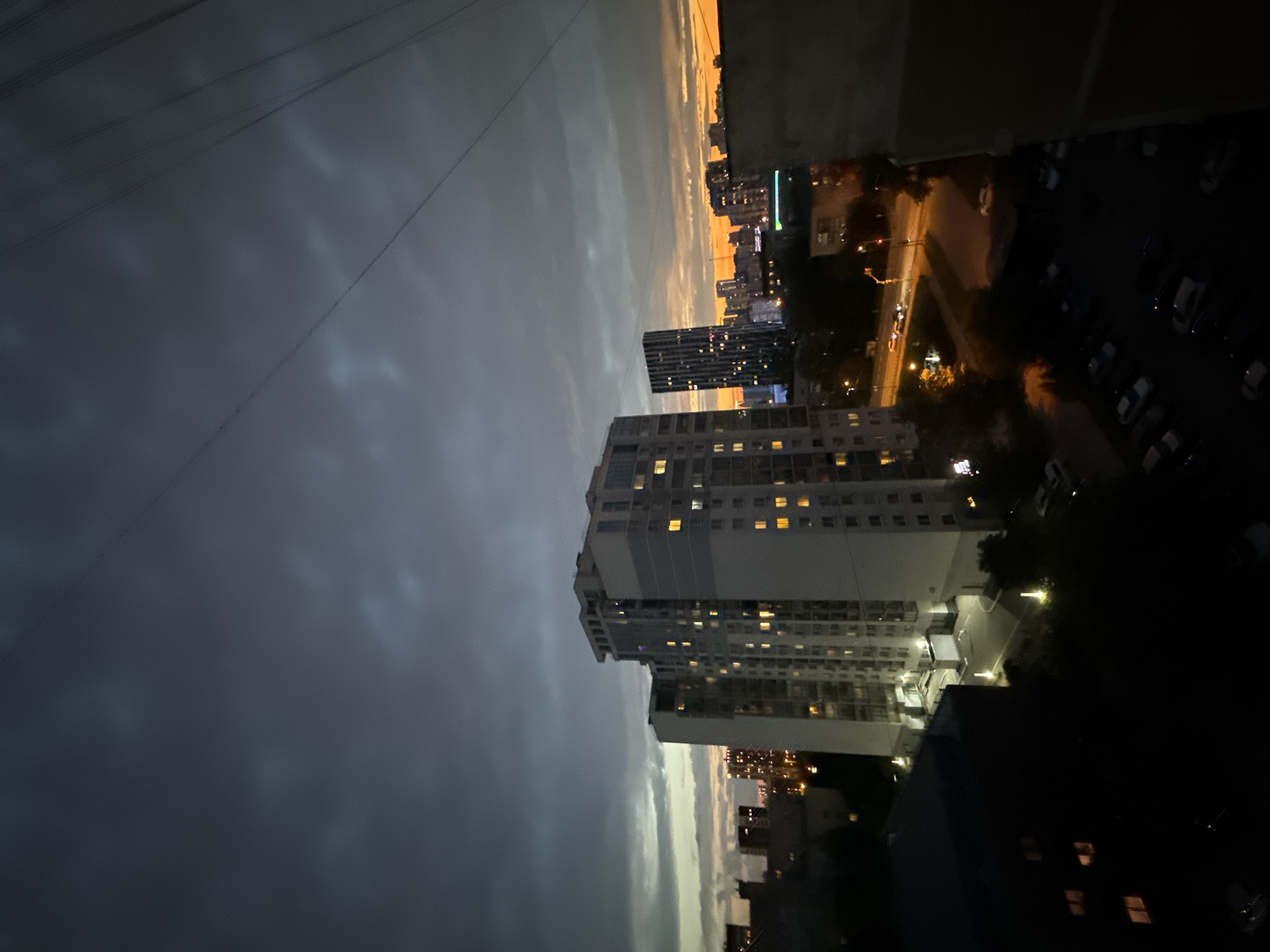 This image depicts a twilight scene in the city of Yekaterinburg, Russia, likely taken from a high vantage point such as an apartment window. The sky is a dramatic mix of dark, cloudy tones in the upper portion and a vibrant orange and yellow sunset glow at the horizon, suggesting it is either early evening or pre-dawn.
Dominating the mid-ground are several multi-story residential buildings. The most prominent is a large, light-colored apartment complex with many windows illuminated, indicating occupancy. To its right, a taller, darker skyscraper with a patterned facade also shows numerous lit windows. Further in the distance, a cluster of city lights from other buildings can be seen against the fading sky.
The foreground is largely dominated by the dark silhouettes of buildings and trees. Below the apartment complexes, streets with cars and streetlights are visible, hinting at ongoing urban activity. A parking lot filled with cars is also discernible in the lower right portion of the image. The presence of power lines crisscrossing the sky further emphasizes the urban environment. No people are clearly visible in the scene. The overall mood is one of a city transitioning from day to night.