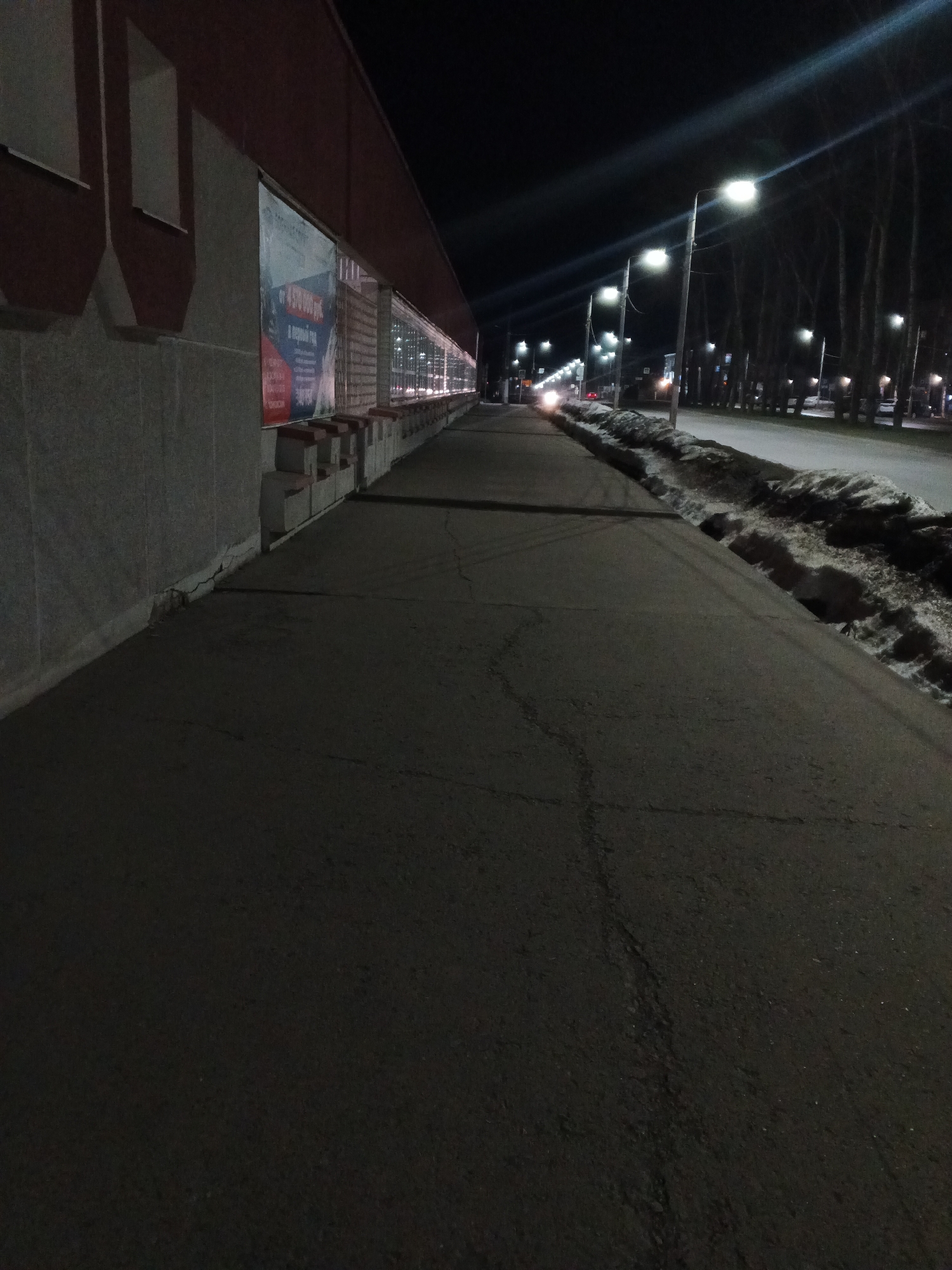 The image depicts a street scene at night in what appears to be the city of Chaikovsky, Russia. Along the left edge of the frame is a building with a light-colored wall and darker red architectural accents. There are signs posted on the building, one of which shows a sum of "4 570 000 руб." A sidewalk runs alongside the building. To the right of the sidewalk, a street with a snowbank alongside it is visible, illuminated by streetlights that line the road. Cars are moving on the street. Trees line the far side of the road, and streetlights continue into the distance. The sky is dark. The sidewalk surface is cracked. The image appears to be taken from a low angle on the sidewalk.