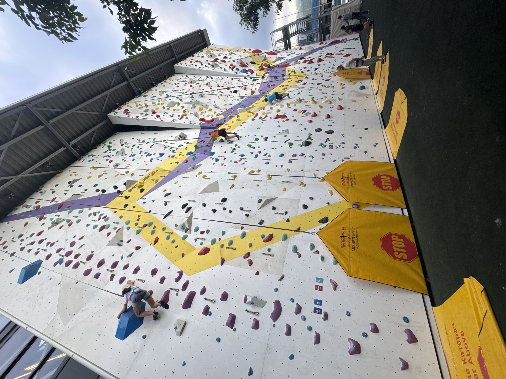 An outdoor rock climbing wall is depicted from a low-angle perspective, showing multiple climbers engaged in the activity under a cloudy sky. The large, white climbing wall is adorned with numerous colorful holds in various shapes and sizes, including red, blue, yellow, green, purple, grey, and black. Distinct climbing routes are marked by broad diagonal stripes of yellow and purple across the wall's surface.
Several individuals are visible on the wall, ascending the routes. In the foreground, a climber in a white and blue top and blue shorts is poised on a large blue block-like hold. Further up, other climbers are seen: one in an orange top, another in a blue top, and a fourth person higher on the wall. At the base of the wall, on a black ground surface, several people are present, one standing near yellow safety mats. A metal roof structure extends over the upper part of the climbing wall, and glimpses of other buildings are visible in the background.
Yellow safety mats or banners are placed at intervals along the bottom of the wall, featuring red "STOP" signs and text in both Bulgarian and English. The Bulgarian text reads "ПРОВЕРЕТЕ!" (Check!) and "ЗАКАЧИХТЕ ЛИ СЕ?" (Are you clipped in?), alongside "DOUBLE CHECK" and "ARE YOU CLIPPED IN?". Another partial phrase visible is "ме Катерач" and "er Above", suggesting instructions related to climber safety. The presence of Bulgarian text confirms the Sofia, Bulgaria location.