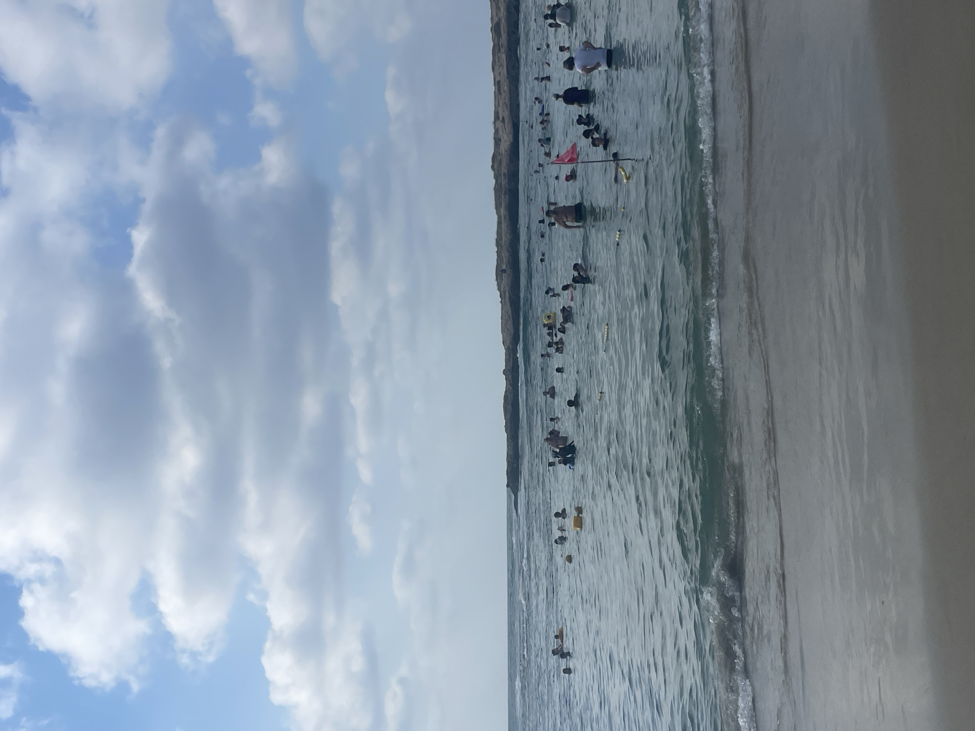 The image depicts a crowded beach with many people swimming in the ocean under a partly cloudy sky. The water is calm with small waves reaching the sandy shore in the foreground. A rocky outcropping is visible in the distance on the right side of the frame, where more people appear to be gathered. Yellow buoys and ropes mark off areas in the water, and a red flag on a pole stands near the center of the scene. The sky is filled with a mix of white and gray clouds. The location is the beach in the city of Hof HaCarmel Regional Council, Israel.